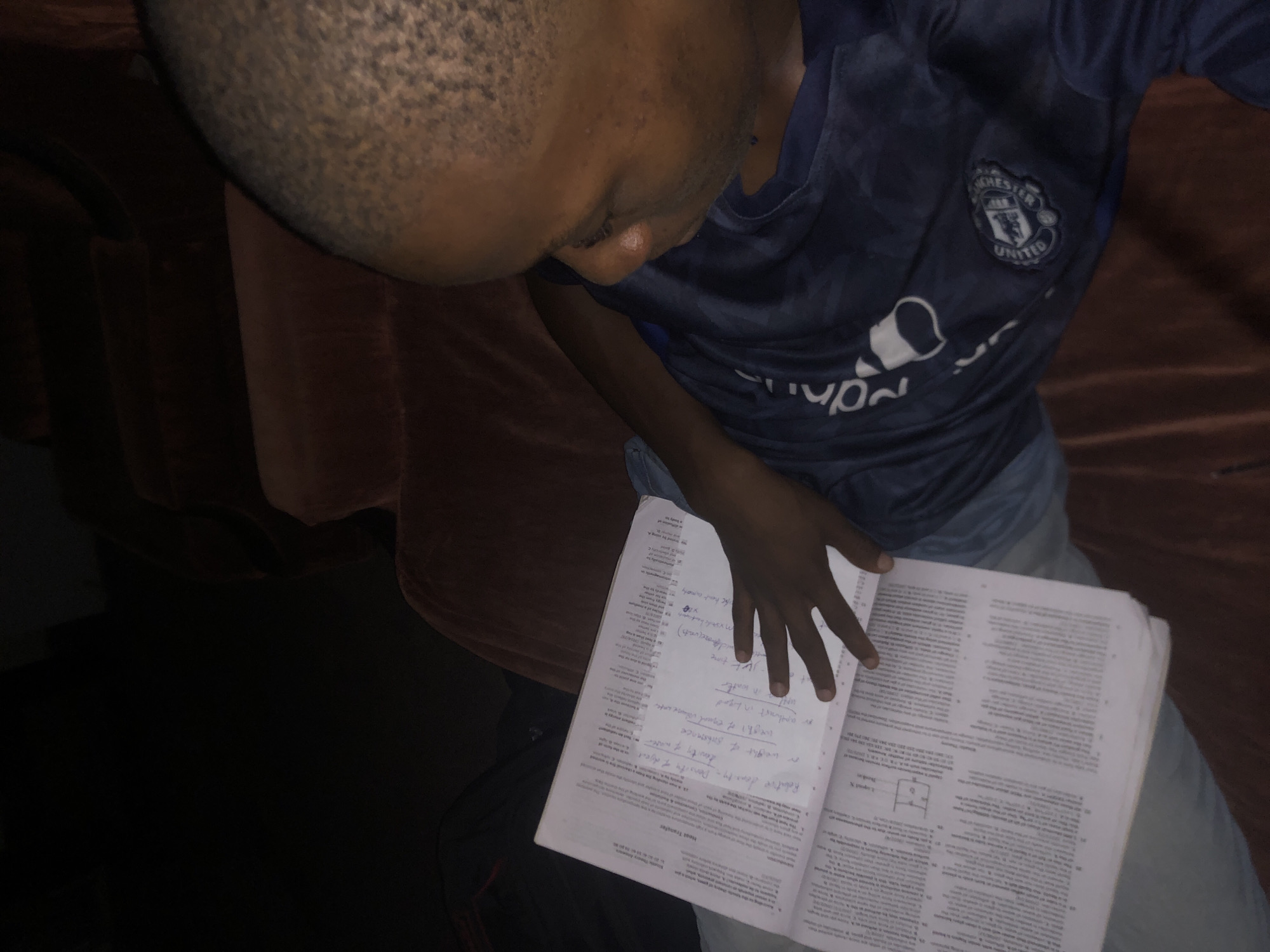 A person with a shaved head, wearing a blue Manchester United shirt and light-colored jeans, is seated on an orange couch and reading a book. The book is open to a page with handwritten notes and printed text about heat transfer and density. The setting appears to be indoors. There is no indication of the city of Atani, Nigeria.