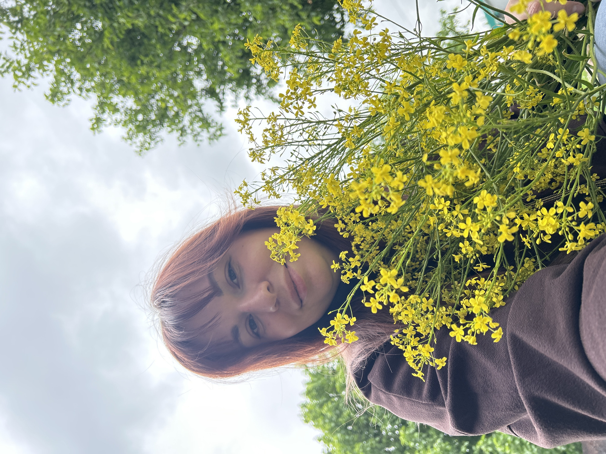 A portrait-style image, rotated 90 degrees counter-clockwise, features a young woman holding a large bouquet of small, bright yellow flowers. Her face, framed by red-brown hair and bangs, is visible on the left side of the frame, looking directly at the camera with a calm expression. The abundant yellow flowers occupy a significant portion of the right side, with some sprigs extending towards her face. She is wearing a dark brown top or jacket. The background, seen from a low angle, includes a light grey, overcast sky and blurred green tree foliage at the bottom left. The image captures an outdoor moment during what appears to be an overcast day. No visible text is present. The scene takes place in Фоки, Russia.