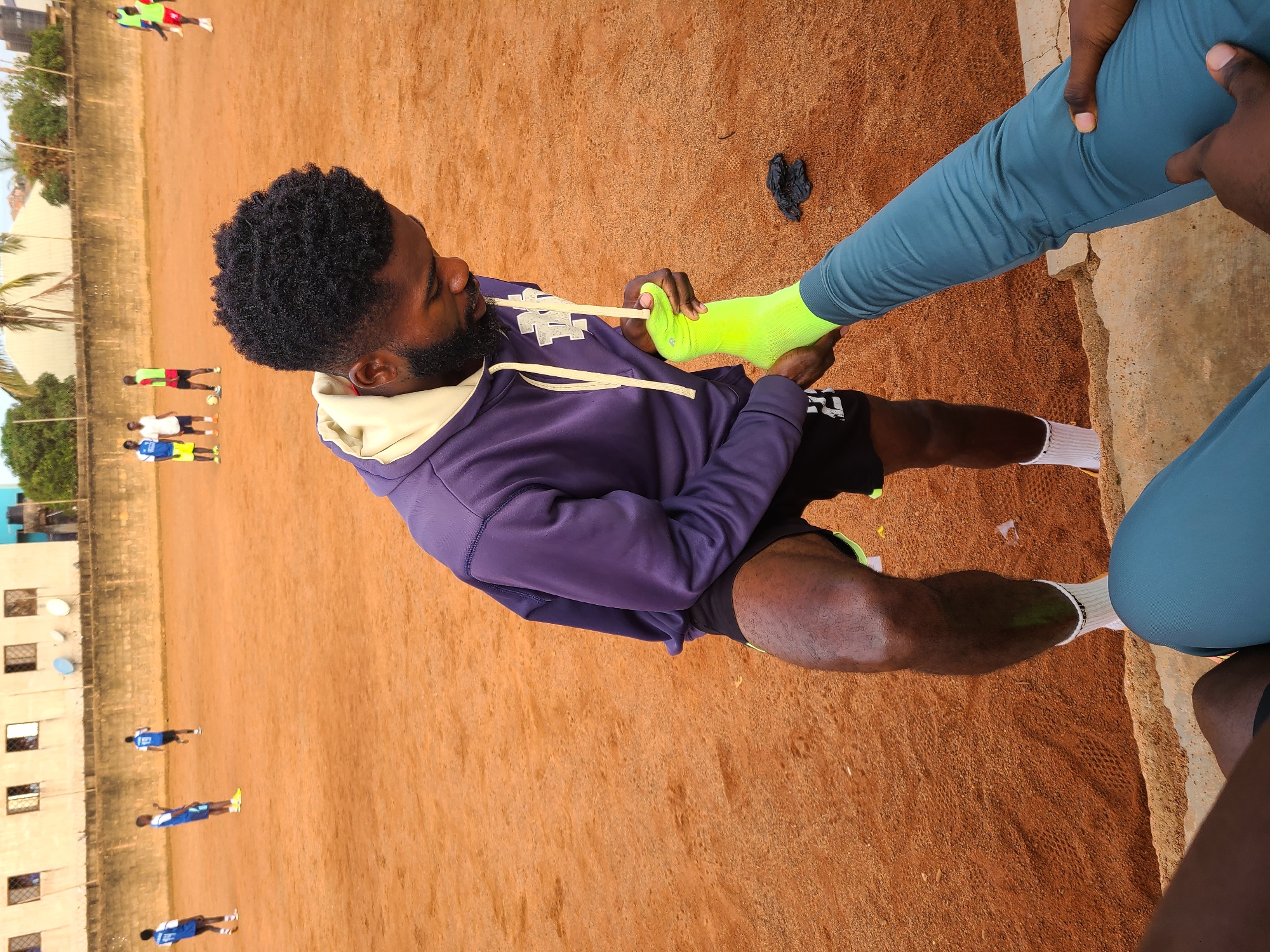 The image captures a young man in Barakin Lerawi, Nigeria, tending to his footwear on a dusty, orange-colored field, likely a soccer pitch. He's dressed in a purple hooded sweatshirt and black shorts. His right leg is raised, and he's pulling on a bright green sock. His other leg is in motion, clad in teal athletic pants. In the background, other individuals are scattered across the field, some appearing to be engaged in a soccer match. Buildings line the perimeter of the field, with some showing signs of wear. The scene suggests a casual sporting event or practice session.