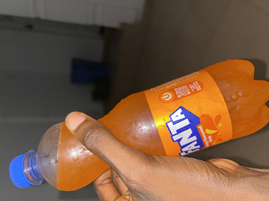 The scene displays a dark-skinned hand holding a clear plastic bottle of Fanta orange-flavored drink. The bottle, covered in condensation, features a blue screw cap and an orange label with the "FANTA" logo in blue and white, "orange flavoured drink" text, a QR code, and the slogan "SIP, SHAKE & SCAN TO UNLOCK THE FUN." The environment is an indoor location in Egbe, Nigeria, characterized by low ambient lighting. The background is dark and out of focus, showing a blurred floor and a distant, dark blue object, consistent with a container.