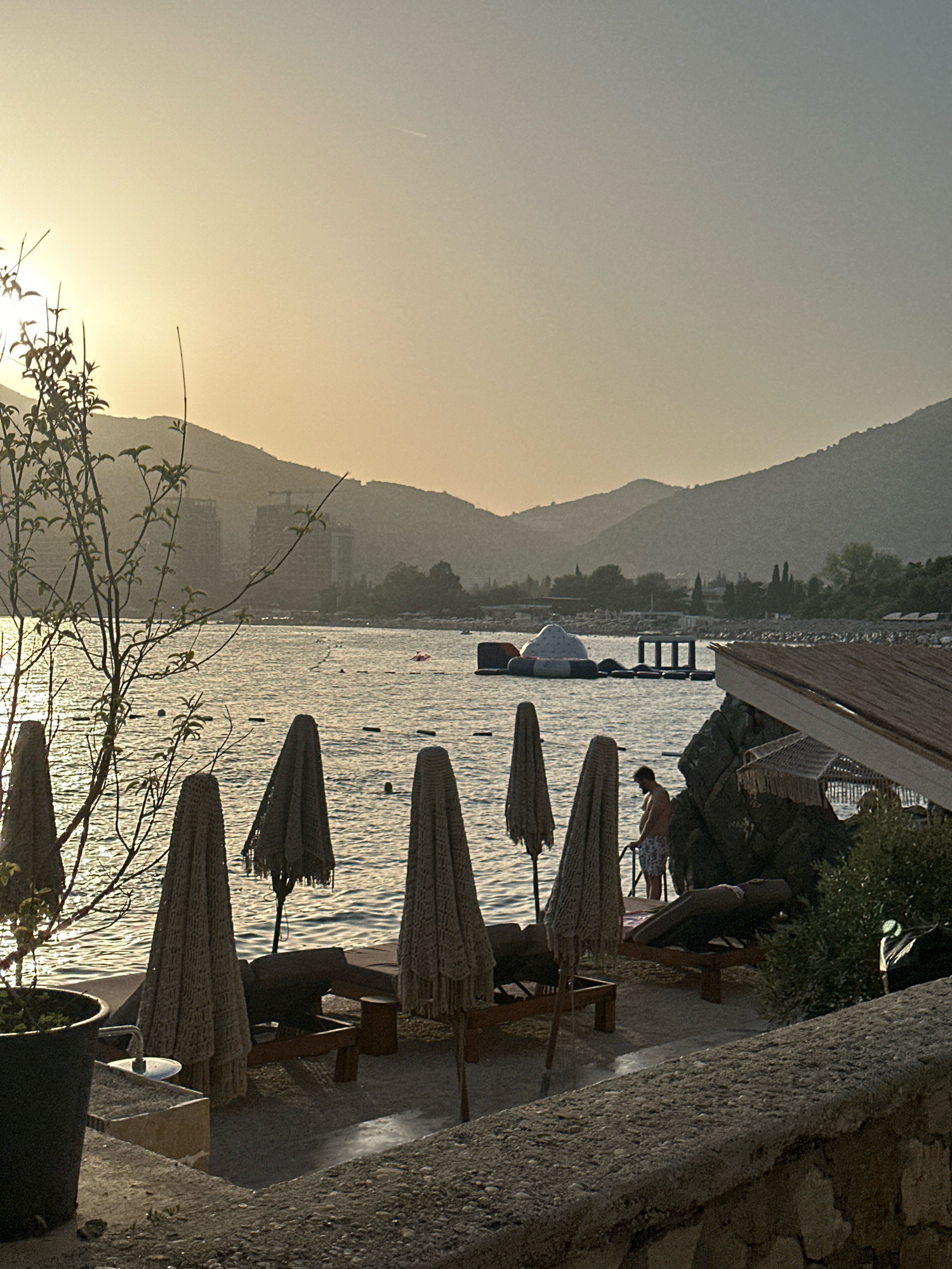 The image depicts a serene beach scene at sunset in Budva, Montenegro. The golden light of the setting sun casts a warm glow over the water and the surrounding hills, creating a peaceful atmosphere. Several lounge chairs and umbrellas are arranged on the sandy shore, indicating a popular tourist spot. In the distance, a man is seen standing near the water, possibly enjoying the evening breeze. A floating inflatable structure is visible in the sea, suggesting recreational activities. The presence of buildings and construction cranes in the background hints at the urban development of the area. The overall composition of the image emphasizes the natural beauty of the coastline, the tranquil ambiance of the beach, and the leisure activities available to visitors.