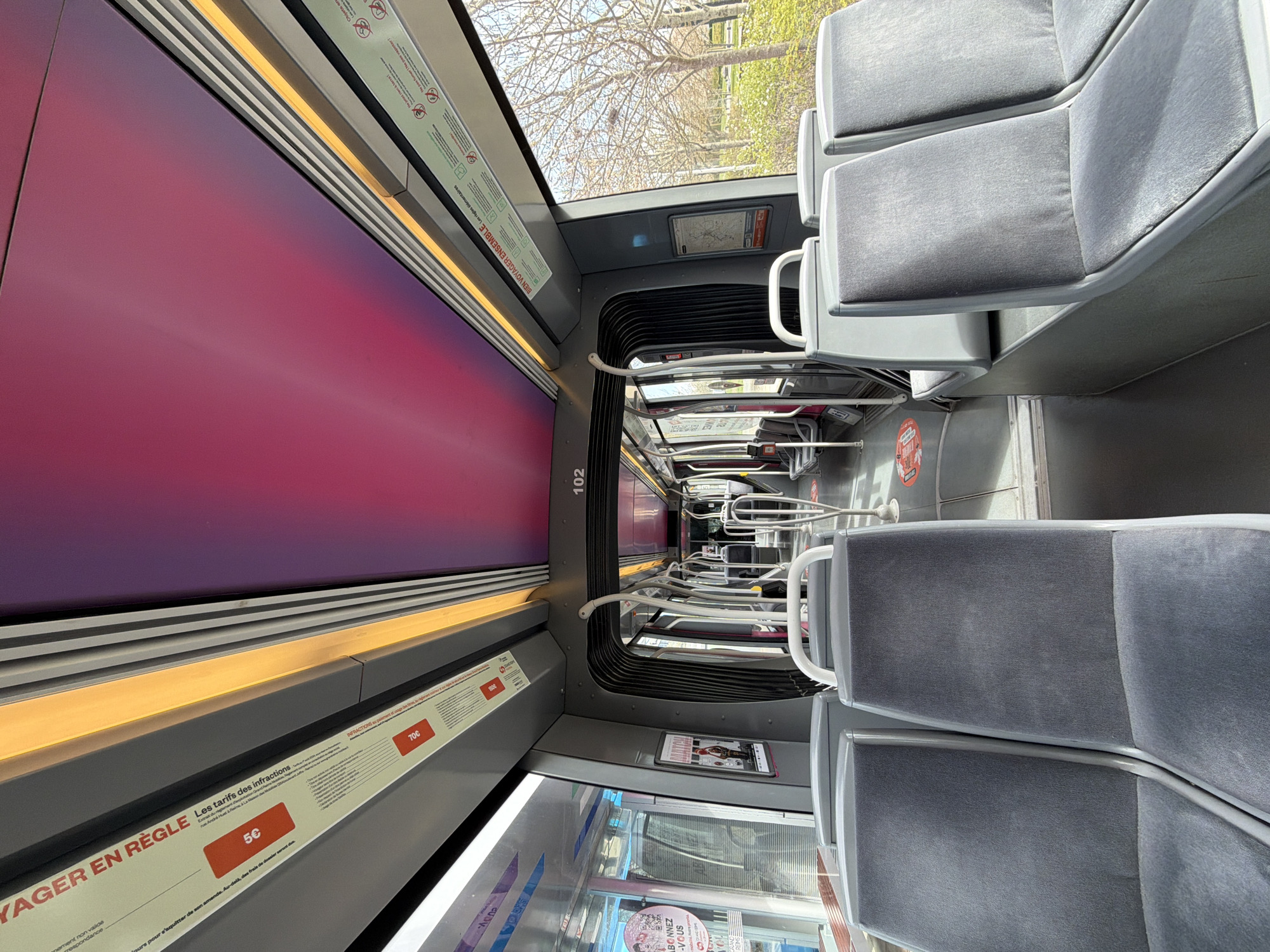 The image depicts the interior of a modern public transport vehicle, likely a tram or light rail, within the city of Les Trois-Domaines, France. The perspective is from the front third of a carriage, looking forward through its length.
The interior is characterized by rows of grey fabric seats with light grey plastic frames, arranged on both sides of a central aisle. The seats in the immediate foreground are unoccupied, with the backs of two pairs of seats visible. The ceiling features a distinctive central panel with a gradient from bright pink to magenta, bordered by grey panels and continuous yellow-orange strip lighting. Grey structural panels and numerous white handrails and stanchions are visible throughout the carriage. A flexible, black accordion-style connector section is present between carriages, visible towards the mid-section.
Various informational signs in French are mounted along the overhead panels. On the left, signs include "VOYAGER EN REGLE" and "Les tarifs des infractions" with associated monetary values "5€" and "70€". On the right, signs include "BIEN VOYAGER ENSEMBLE" accompanied by pictograms indicating regulations (e.g., no food, no smoking). A digital display further down the carriage shows the number "102". A framed poster featuring "MATCH" and a person is affixed to a side panel on the left, while a route map is visible on a panel to the right. The floor is dark grey, and red circular adhesive markings with white footprints are partially visible on the aisle floor, indicating designated standing areas.
One person is faintly visible sitting in a seat on the right side of the carriage, further down, largely obscured by the interior structure. The majority of seats appear empty.
Through the windows on the right, an outdoor scene is visible under bright daylight, showing bare deciduous trees with their branches prominent, green ground cover, and a clear blue sky, indicative of late winter or early spring. The windows on the left reflect parts of the interior and show glimpses of an urban environment outside.