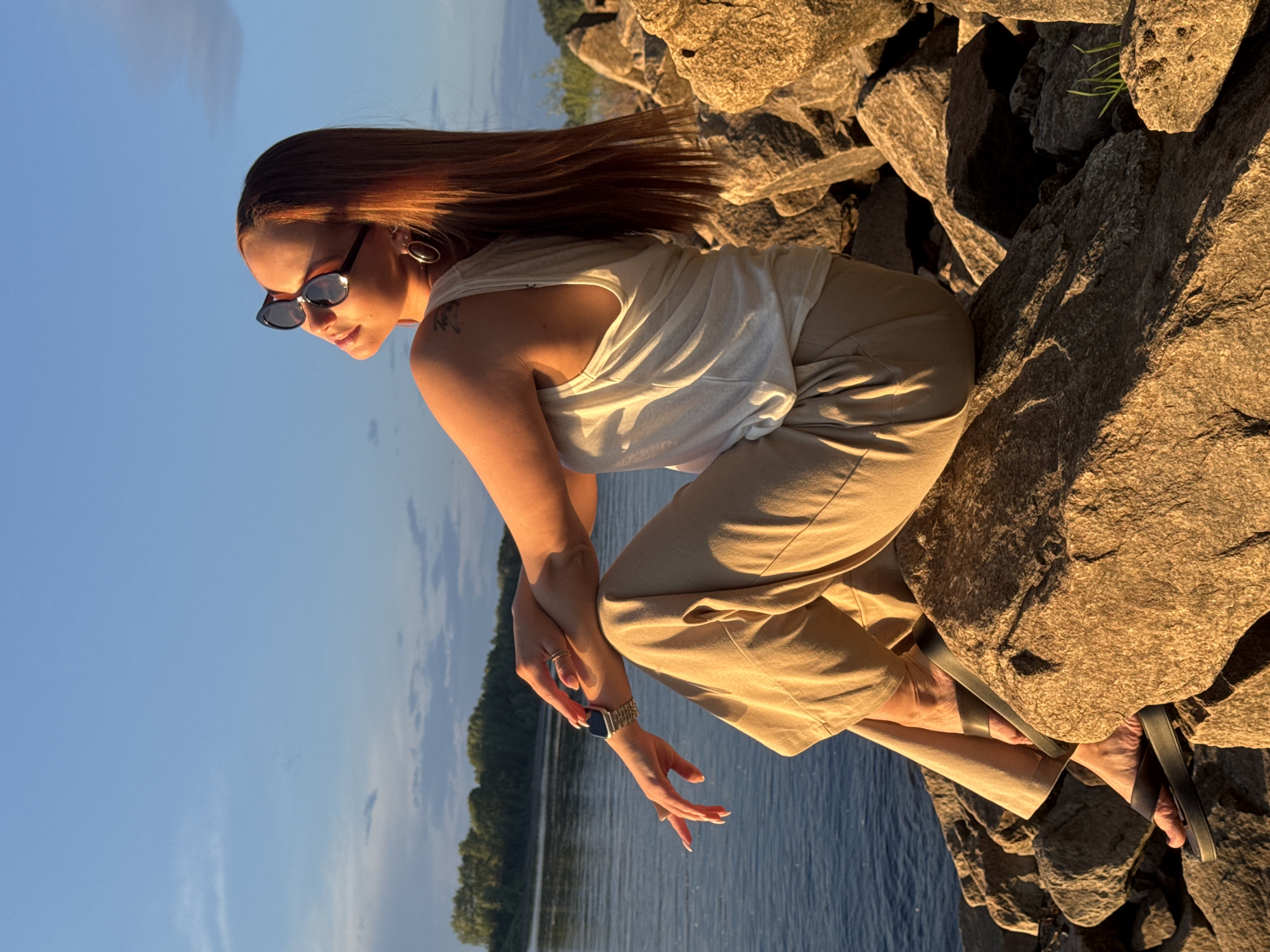 In this outdoor, eye-level shot, a young woman is captured in a side profile, seated on large, rough rocks. The setting appears to be a natural landscape, with a body of water and a treeline visible in the background, suggesting a riverside or lakeside location. The time of day is likely late afternoon or early evening, as indicated by the warm, golden light illuminating the scene and casting long shadows. The sky is a clear blue with a few wispy clouds.
The woman has long, reddish-brown hair that cascades around her shoulders. She is wearing a white tank top and light-colored, loose-fitting pants. Her eyes are hidden behind black sunglasses. She is adorned with a silver hoop earring in her visible ear and a watch on her left wrist. Her hands are gently clasped together, resting on her knees, with her fingers slightly splayed. Her posture suggests a relaxed and contemplative mood, possibly enjoying the serene environment. She is also wearing dark flip-flops.
The rocks she is sitting on are substantial and textured, with some patches of greenery growing between them. The water is a deep blue and appears calm. The overall atmosphere is peaceful and tranquil, bathed in the soft glow of the setting sun. No text is visible in the image.