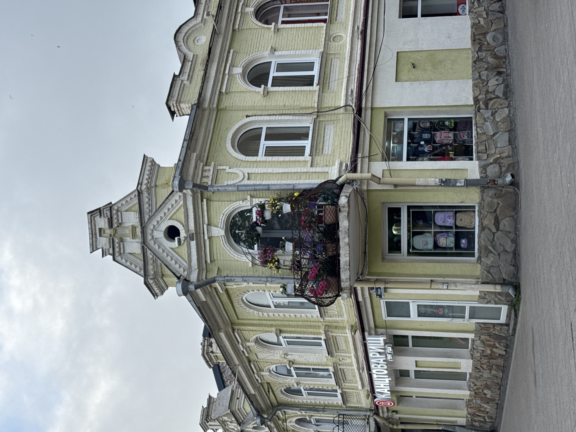 The image depicts a multi-story building in Akhieli, Georgia. The building features an ornate facade with arched windows, a small balcony adorned with flowers, and a decorative gable. The exterior is painted in light yellow and white. A sign in Cyrillic script, “Канцтоварищ (since 1997 years),” indicates a stationery store at the ground level. The windows on the ground floor display merchandise, including backpacks. The building is situated alongside a paved street. The sky is overcast.