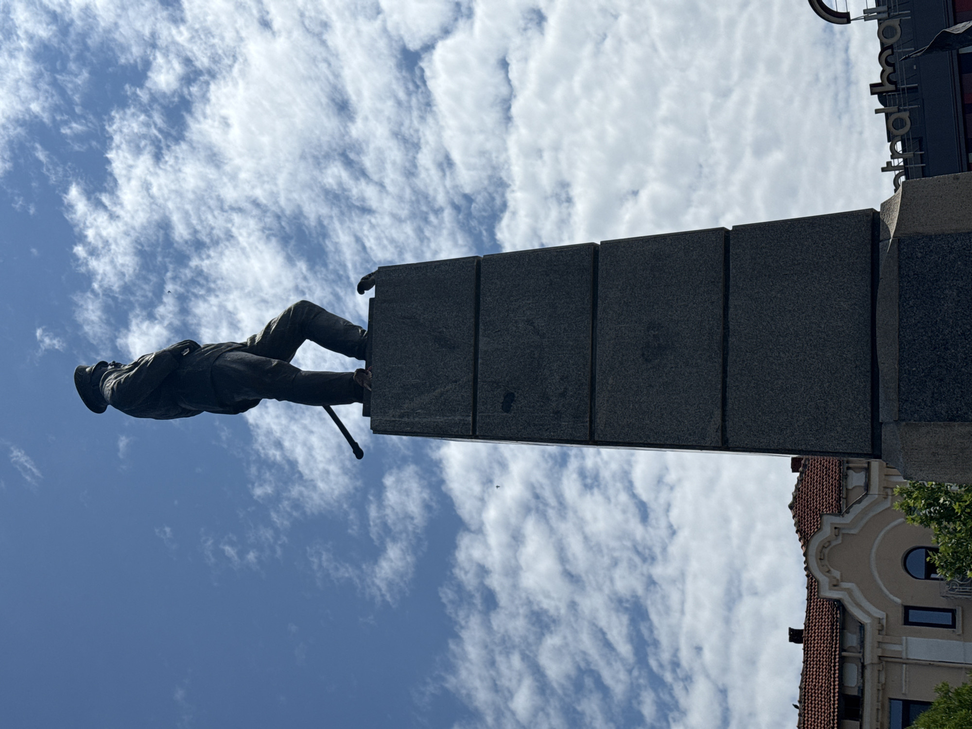 The original image is presented on its side, but for clarity, this description assumes a corrected vertical orientation.
The central subject is a majestic dark-colored statue of a man, likely cast in bronze, standing atop a tall, imposing pedestal. The figure wears a formal outfit, including a hat, a long coat, and trousers, and holds a walking stick or cane in his left hand. His stance is dignified, looking forward or slightly upwards. The pedestal is constructed from dark gray, polished stone blocks, giving it a solid and monumental appearance.
The background consists predominantly of a bright blue sky dotted with scattered, wispy white clouds, suggesting a clear and sunny day. In the upper right corner, parts of urban architecture are visible: a building with a distinctive red-tiled roof and an ornate, light-colored facade featuring decorative moldings and windows. Above this building, a partial sign displays upside-down letters that appear to spell out "palombaro" or a similar word, suggesting a commercial establishment. A tiny dark speck, possibly a distant bird or aircraft, is visible in the expansive sky.
The photograph is taken from a low vantage point, looking up at the statue and the sky, which enhances the statue's imposing presence. The scene captures a public monument in what appears to be a European town, consistent with the given context of Pleven, Bulgaria.