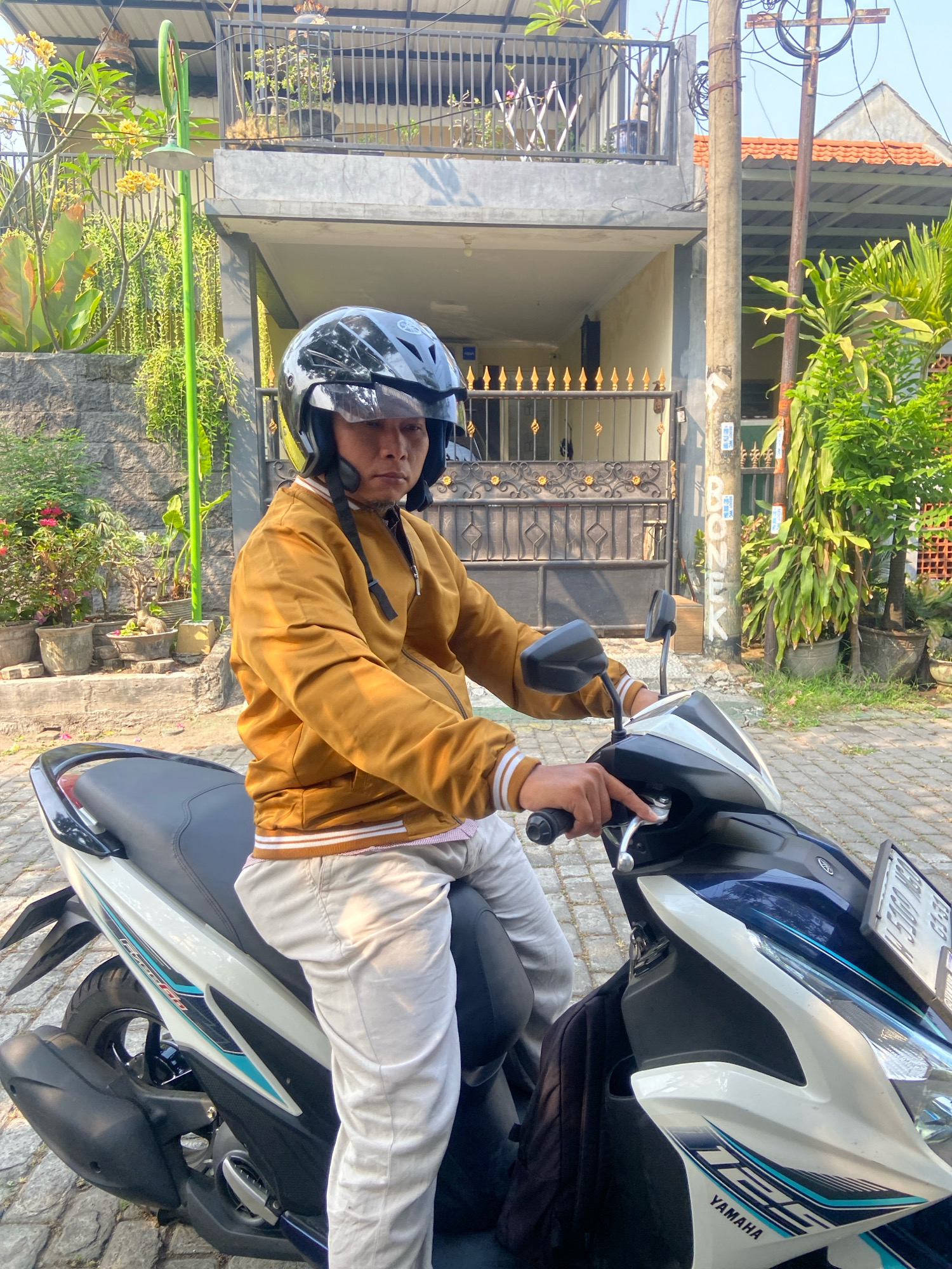 A man wearing a brown jacket and light-colored pants is sitting on a blue and white motorcycle. He is wearing a black helmet with a clear visor. The man is in Gelam, Indonesia. The background shows a house with a metal gate and several potted plants. The day appears to be sunny.