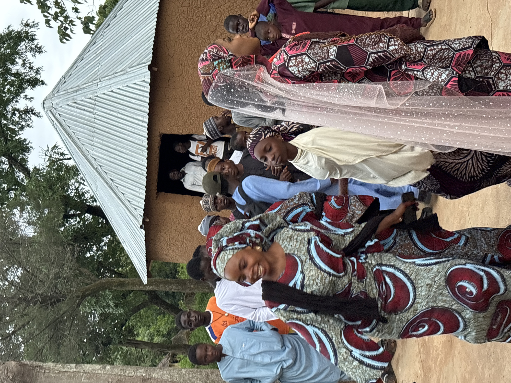 This media file shows a gathering of people outside a building with a corrugated metal roof and a mud-brick wall. The subjects are predominantly African individuals, many of whom are dressed in vibrant, patterned fabrics. The overall mood appears celebratory or social, with several people smiling and engaged with each other.
One woman in the foreground is wearing a colorful dress with a red, blue, and white swirling pattern and a matching headscarf. She is laughing and appears to be the focus of some attention. Beside her, a person in a light blue garment is looking towards the camera. Other individuals are visible in the background, some peering out from a doorway, and others standing around the gathering. One person is draped in a light pink, sheer fabric with white polka dots, possibly a veil or celebratory attire. The clothing styles suggest a cultural event, possibly a wedding or a similar ceremony.
The scene is outdoors, with trees and foliage visible on the left side of the image, indicating a rural or semi-rural setting. The lighting suggests it is daytime, likely overcast or nearing evening due to the diffused light. The presence of a mud-brick building and the attire of the people are consistent with a location in Jos, Nigeria. No visible text is present in the image.