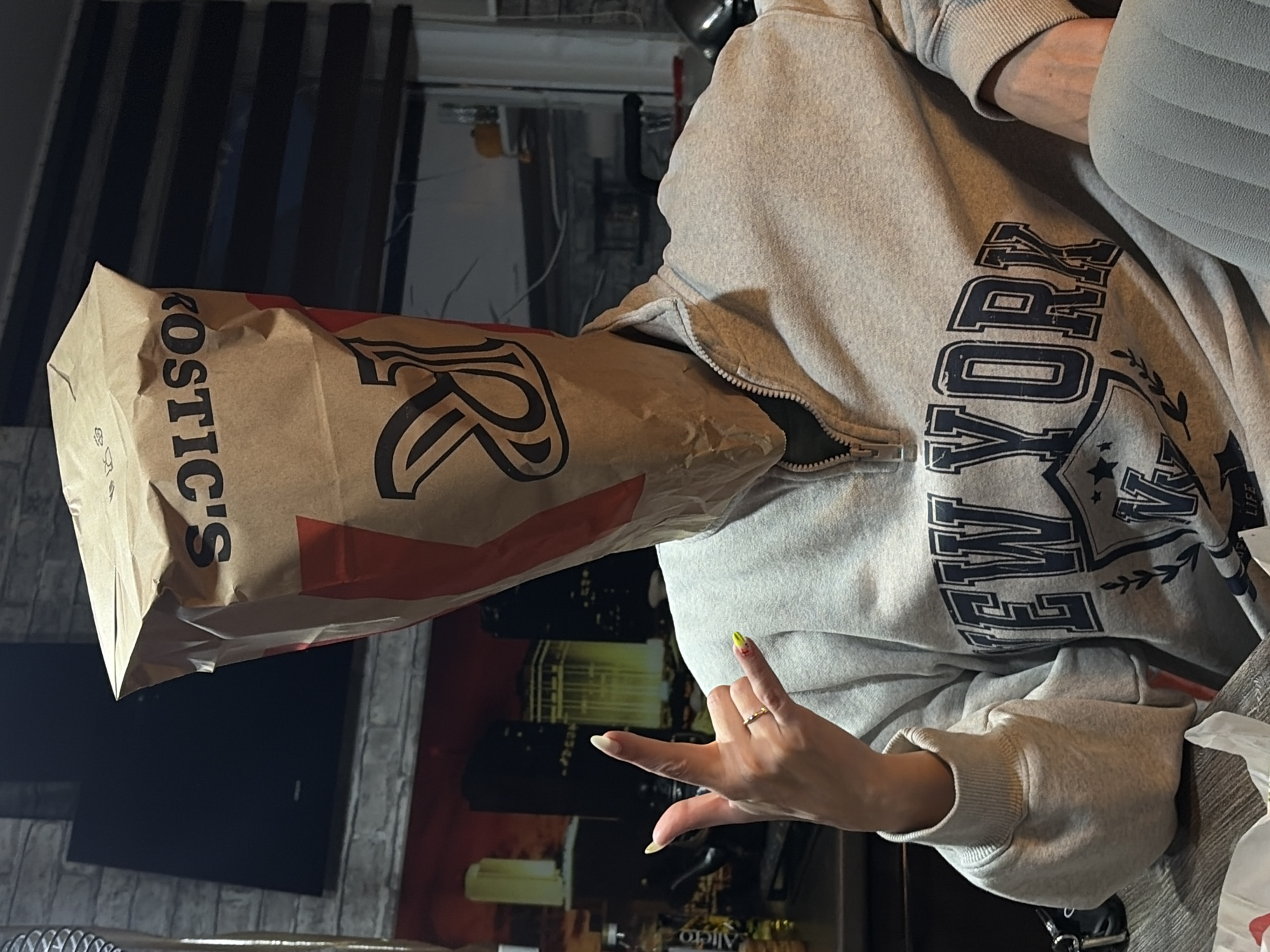 A person is wearing a gray hoodie that reads "NEW YORK" and has a paper bag on their head. The bag has the word "ROSTICS" on it with a red logo. The person is holding up their left hand with their index and pinky fingers extended. They are wearing a gold ring on their ring finger. The background includes a television and a wall decoration depicting a city skyline at night. The city's name and state are unknown.