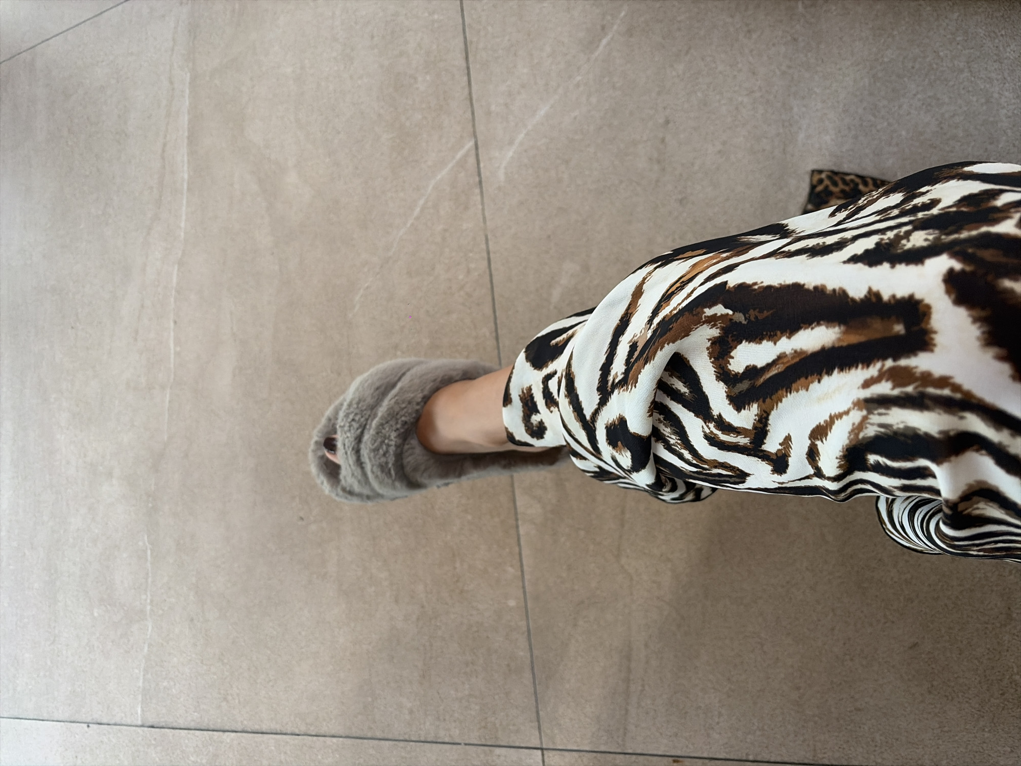 In North Kuta, Indonesia, a person's left leg, clad in a fuzzy grey slipper and patterned pants, rests on a tiled floor. The pants exhibit a bold black and white zebra print. The tiled floor is a light beige with visible grout lines.