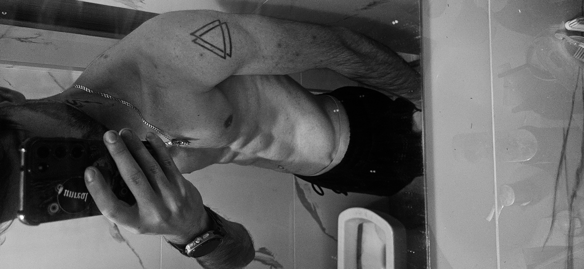 A black and white photograph depicts a man taking a selfie in a bathroom mirror. The man is shirtless, wearing black shorts, a watch, and a chain necklace. He has a tattoo of two intersecting triangles on his left shoulder. The bathroom features a marble-like countertop, a white sink, and toiletries visible in the mirror's reflection. The photograph's orientation is rotated, and there is no indication of the location being in Plovdiv, Bulgaria.