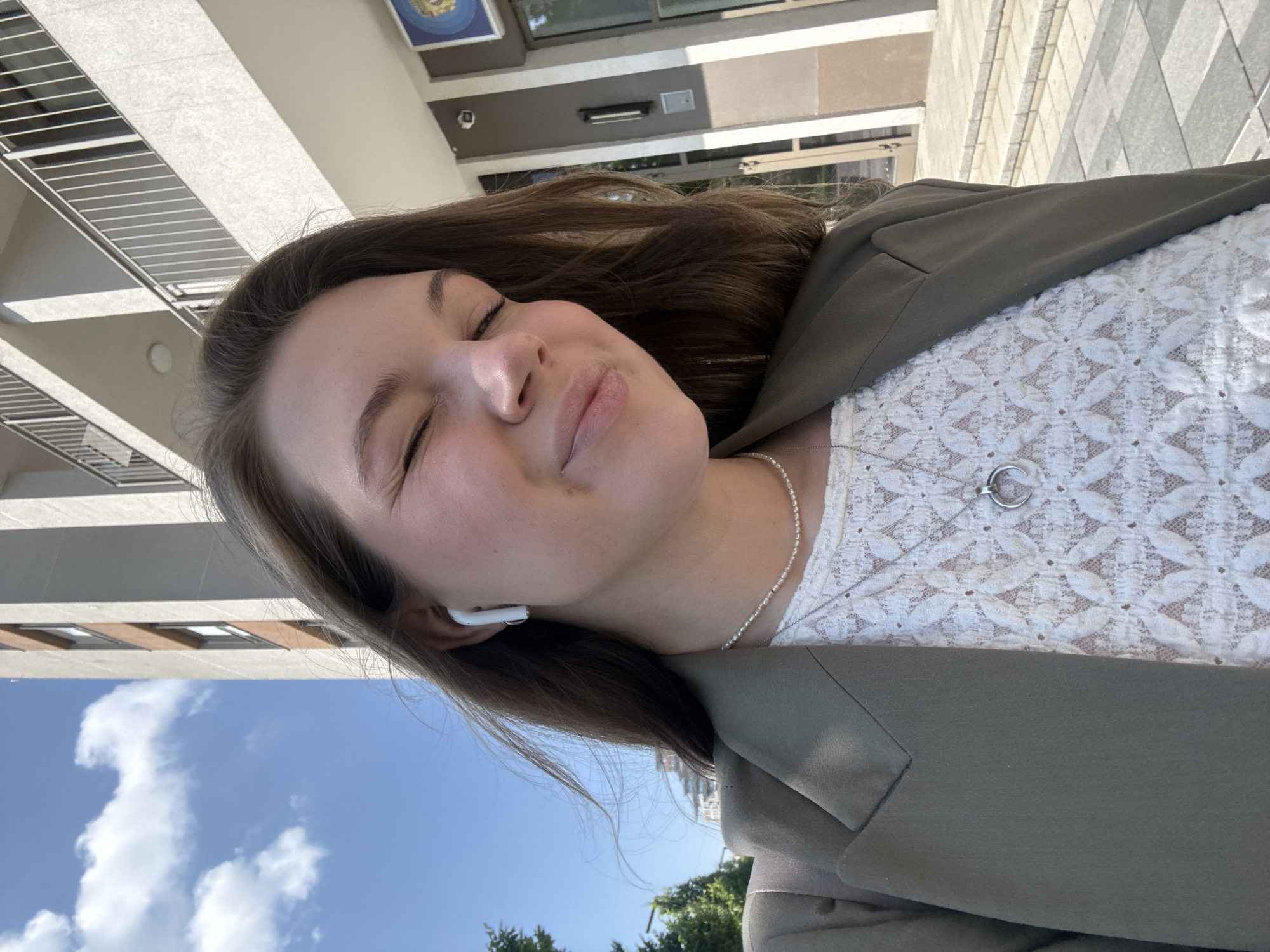 A woman with brown hair wearing a white lace top and a green blazer is outdoors, likely in an urban environment. She has her eyes closed and appears to be smiling slightly. She has an earpiece in one ear. She is also wearing a silver chain with a pendant.
Buildings and a cloudy sky are visible in the background. Based on the architecture and building design visible in the frame, as well as the type of street paving, it is highly probable that the location is in the city of Belgrade, Serbia.