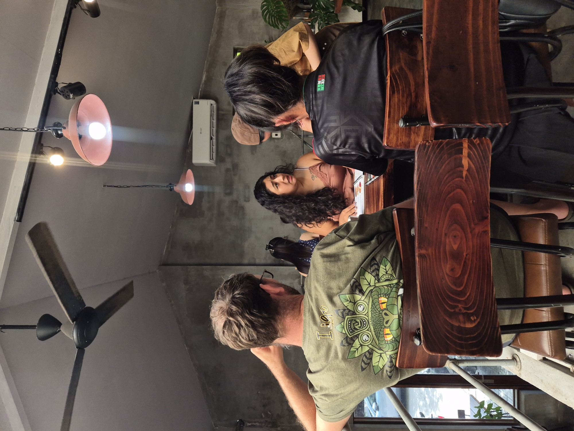 In a modern cafe in Da Nang, Vietnam, with concrete walls and industrial-style lighting, a group of patrons are seated at wooden tables. In the foreground, two individuals are viewed from behind, one wearing a green t-shirt with a graphic of a frog-like creature, and the other in a black shirt featuring the Mexican flag. A woman with long, dark curly hair is seated at a table, engaged in conversation with another person whose face is partially obscured. The cafe is furnished with simple wooden chairs and tables, and a ceiling fan is visible overhead. The overall ambiance suggests a casual gathering or meal.