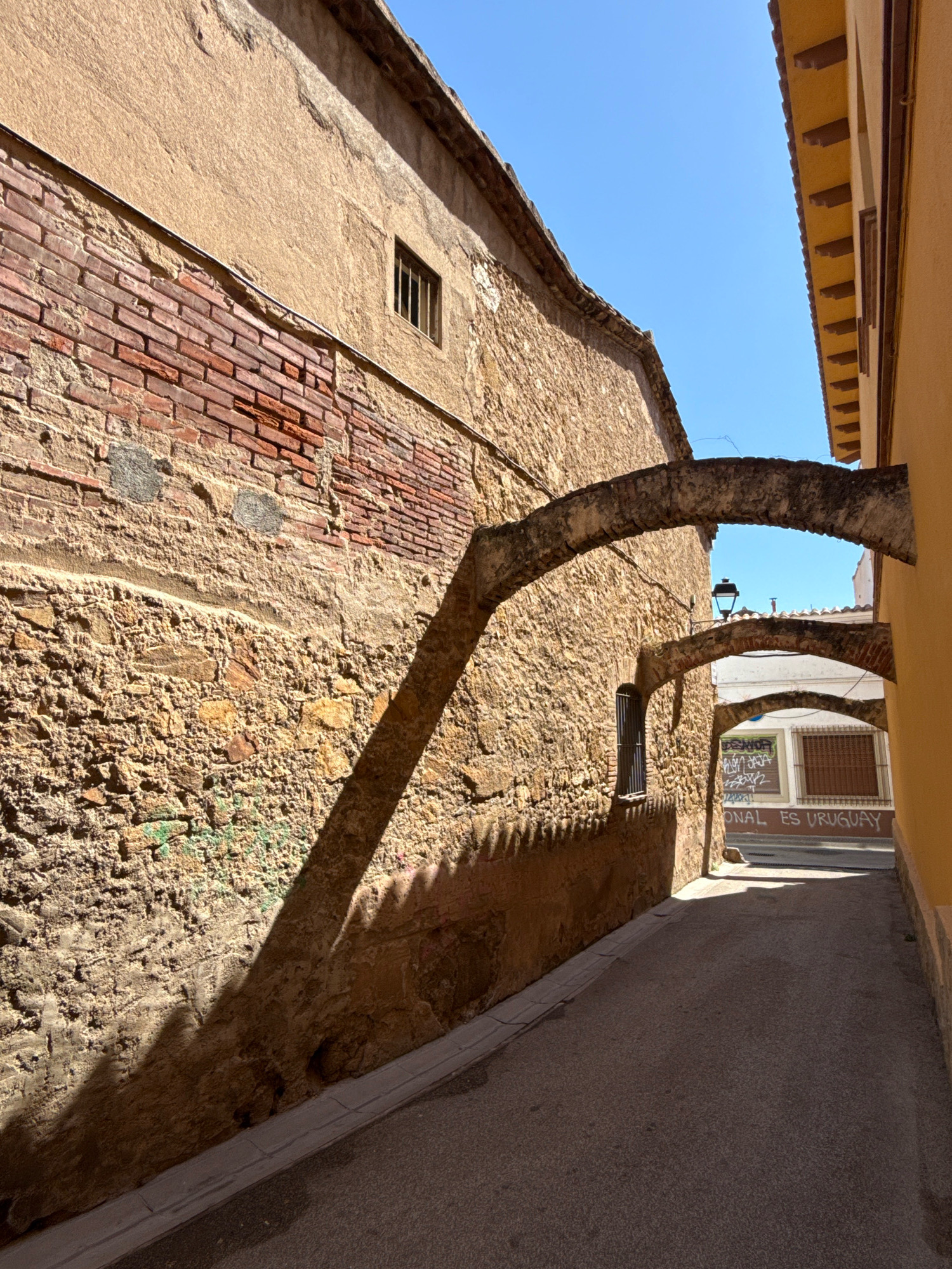 This image shows a narrow street in Malgrat de Mar, Spain, lined with old stone buildings. Sunlight streams down, casting long shadows from arched passageways that span the street. One building features a weathered facade of rough stone and exposed red brick, with a small, barred window. On the opposite side of the street, another building displays a bright yellow exterior. Further down the street, graffiti is visible on a wall, with the words "ONAL ES URUGUAY" painted in red. The sky is a clear, bright blue, indicating a sunny day. The scene captures a quiet, historic corner of the town.