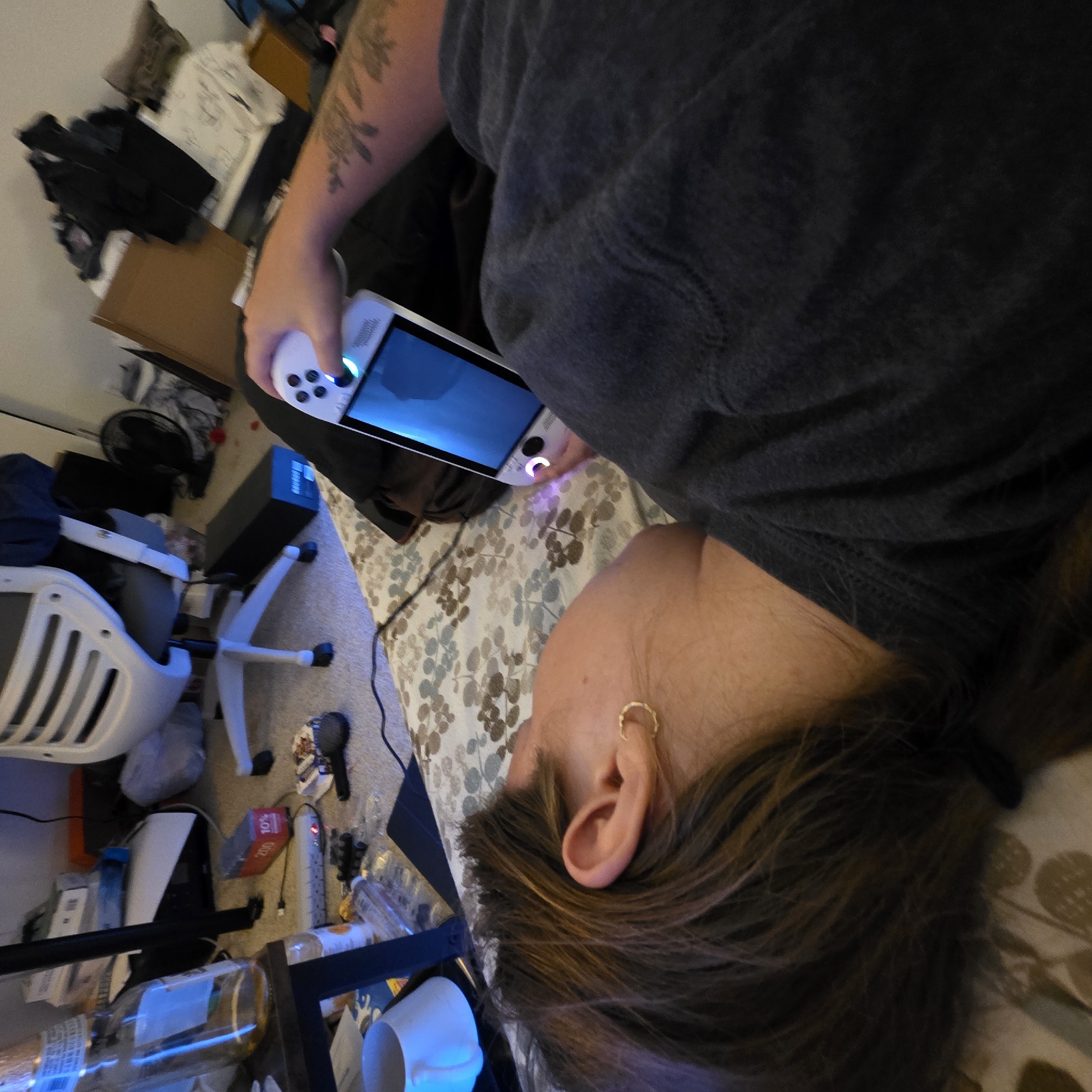 The image shows a person lying on a bed, playing a handheld gaming device. The room appears to be a bedroom or dorm room, with a desk, chair, and various other objects scattered around. The person is wearing a dark t-shirt and has long brown hair and a tattoo on their arm. The gaming device is white and has a large screen. The overall impression is that of a casual, relaxed setting. It is not possible to determine the city of Portland or the United States of America from the image.