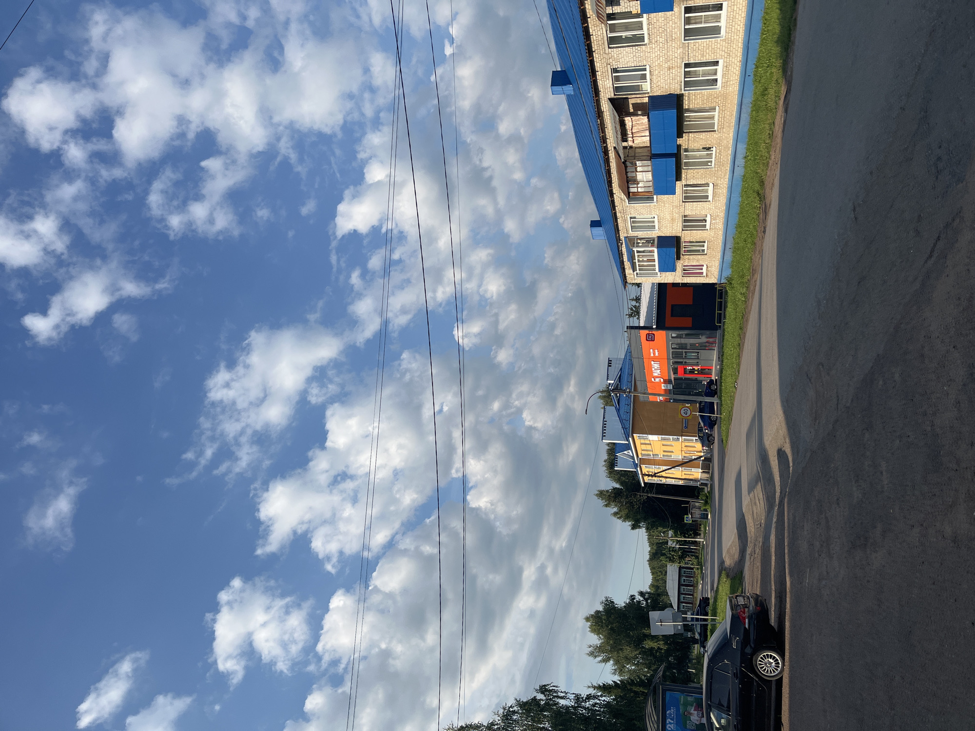 This is a photograph of a street scene in Blagoveshchensk, Russia, likely in a residential or mixed-use area. The perspective is from the side of the road, looking forward.
In the foreground, there is an asphalt road with a shadow cast across it, suggesting it is late afternoon or early morning. A dark-colored sedan is parked on the left side of the road.
Along the right side of the street, there is a light-colored brick building with a bright blue roof and blue balconies. This building appears to be a multi-story apartment complex. Next to it, to the left, is a modern-looking commercial building with orange and blue signage. The visible signage includes "МАГНИТ," which is a well-known Russian supermarket chain.
Across the street, further in the distance, are other buildings, including a yellow building and some smaller, more traditional-looking houses. There are trees lining the street and in the background. Overhead, multiple power lines stretch across the sky.
The sky is a vibrant blue with scattered white clouds. The overall impression is of a daytime scene in a moderately developed urban or suburban environment. There are no people visible in the photograph. The environment appears to be clean and well-maintained.