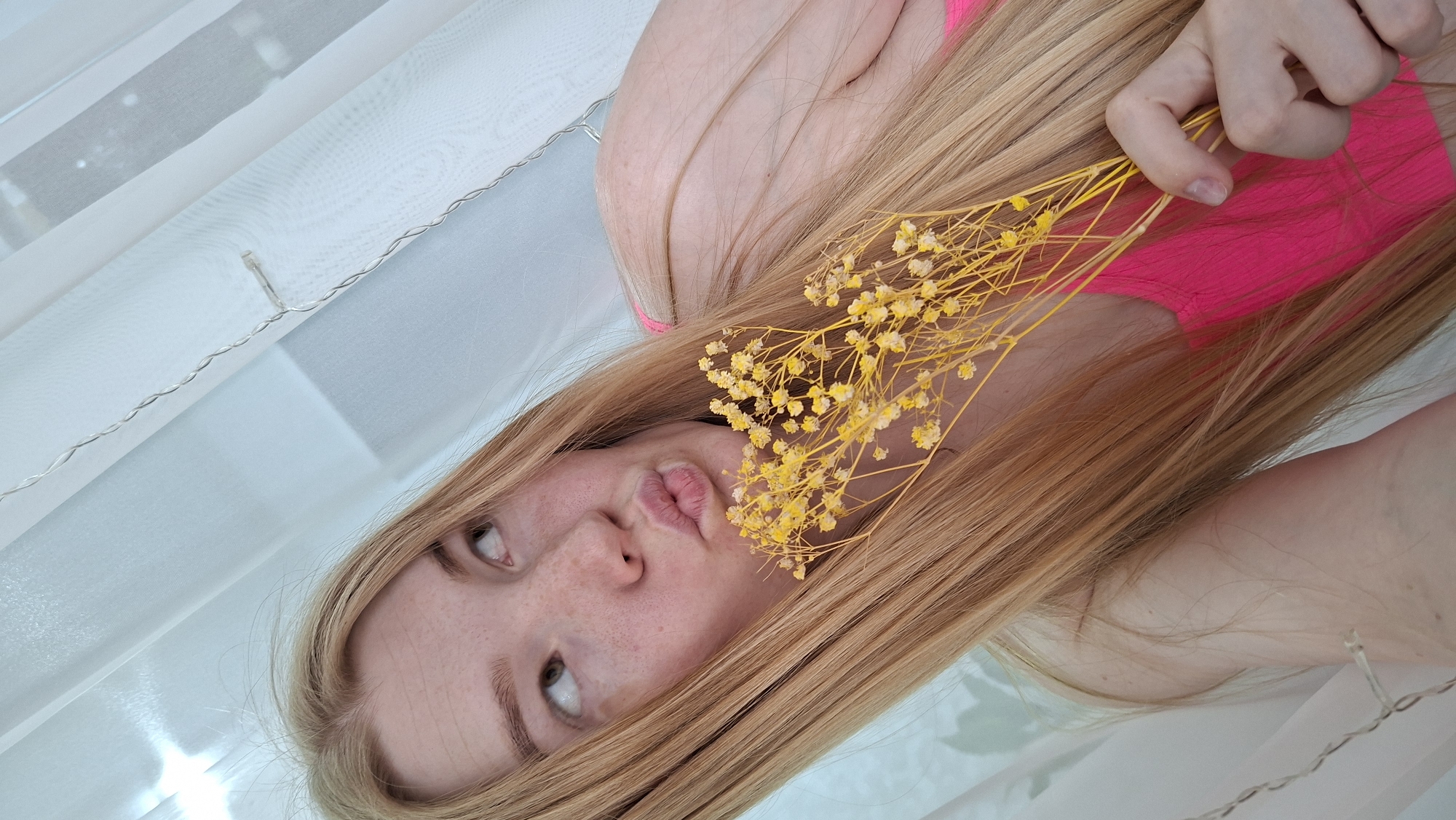 A young fair-skinned person with long, straight blonde hair is angled in the frame, holding a cluster of small, dried yellow flowers near their face, which has a slightly puckered expression. The person is wearing a bright pink top. The background appears to be a soft, white fabric or curtain, and there's a faint string of lights visible in the bottom corner and along the top. The location is listed as Chaikovsky, Russia, but there is no information in the image to support or deny this.