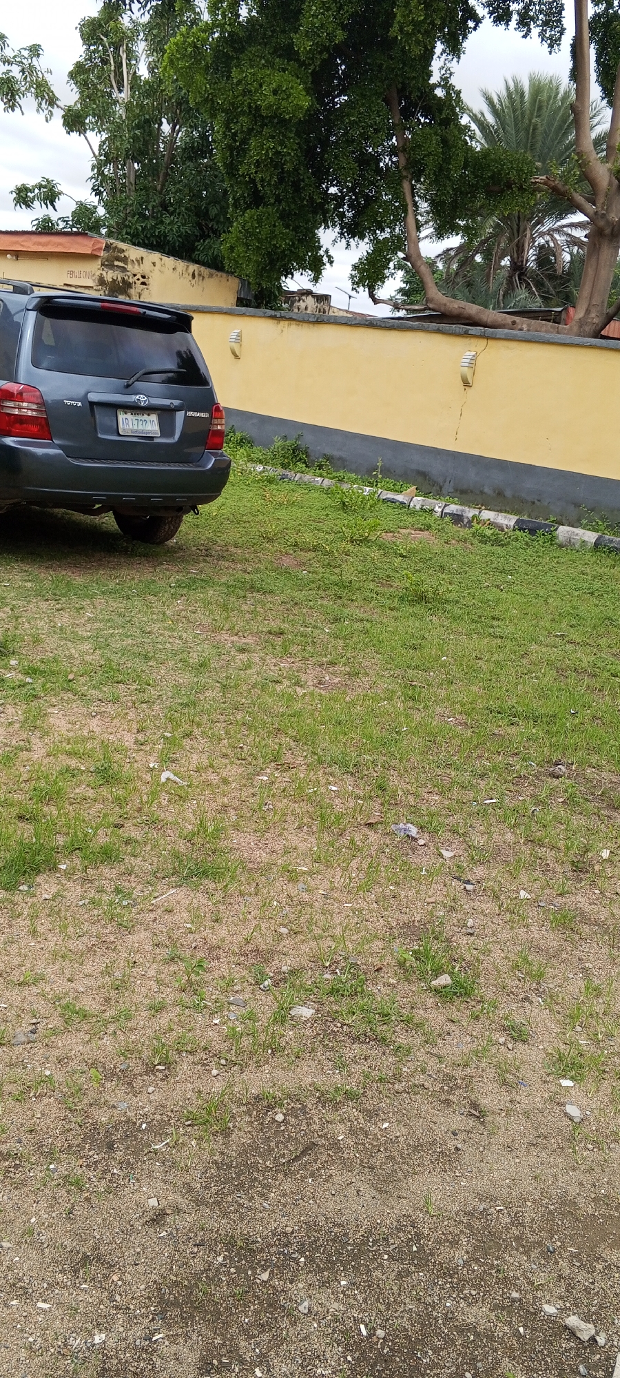 The image depicts the rear of a dark grey Toyota Highlander SUV parked on a patch of grass and dirt. The vehicle's license plate is visible, reading "ABJ 732 JD". Behind the car, a yellow wall runs horizontally across the frame, with two outdoor lights mounted on it. Several trees, including a large leafy tree and a palm tree, are visible behind the wall. The ground in the foreground is a mixture of sand and short green grass, with some scattered debris. The scene appears to be an outdoor area in Jalingo, Nigeria.