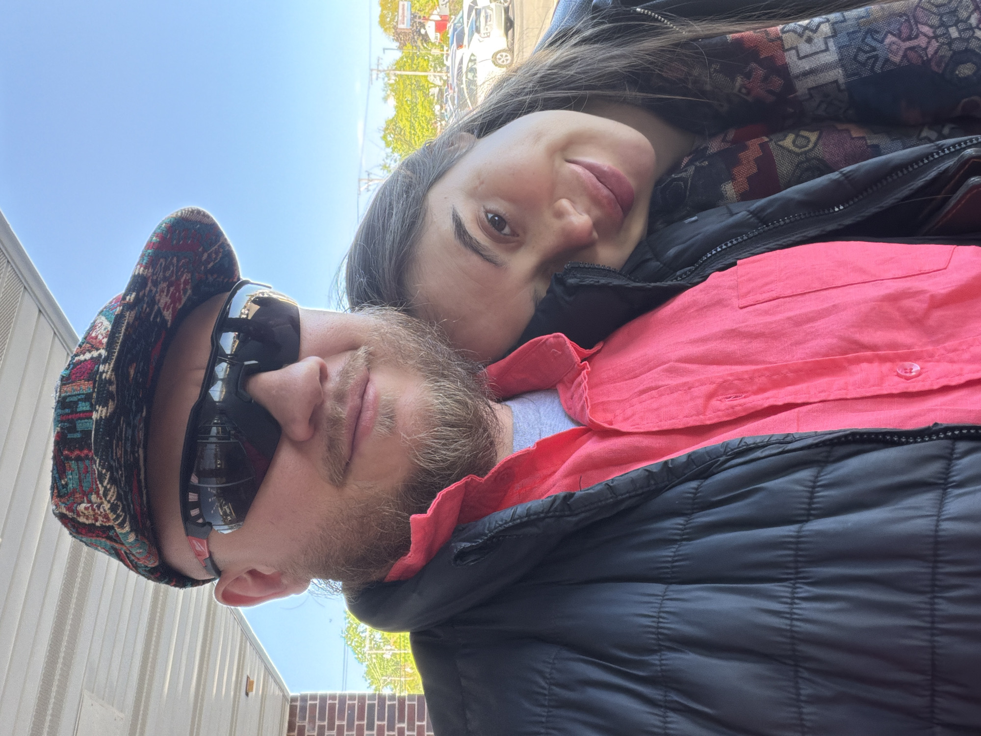 A photograph depicts a man and a woman posing closely, with the woman leaning her head on the man's chest. The man wears sunglasses and a patterned cap. He also has a beard. He is wearing a red shirt with a black puffer jacket on top. The woman has long brown hair and is wearing a patterned scarf and jacket. The image appears to have been taken outdoors. Behind the couple, partially obscured by trees, are parked vehicles and buildings. The sky is visible in the upper left corner of the frame. The location is Niles, United States of America.