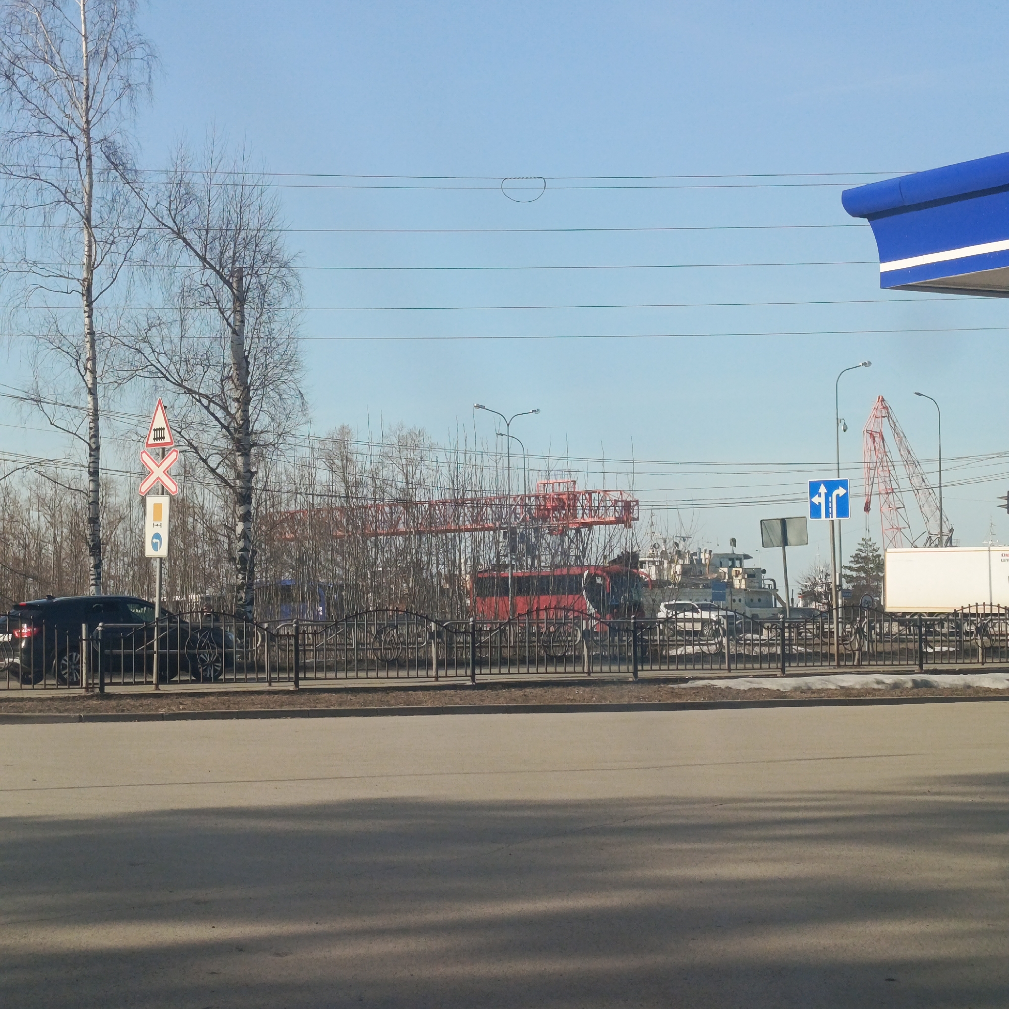 The image depicts a street scene in what appears to be the city of Chaikovsky, Russia, based on the signage, typical urban landscape, and Cyrillic script potentially visible on signs. The foreground consists of a paved street with a substantial area covered in shadow. Beyond the street is a low, black metal fence separating the street from what appears to be a service road or parking area adjacent to a more complex industrial or commercial area.
In the middle ground, behind the fence, several objects are visible, including cars, trucks, and a large red crane with a prominent boom. A bus and numerous vehicles are partially obscured by trees and the fence. Road signs are posted, including a "railroad crossing" sign and a direction sign with a blue background and white arrows. A gas station or commercial building is visible on the right edge of the image, indicated by a blue roof overhang.
In the background, numerous buildings and industrial structures are visible, suggesting an industrial area, possibly a port or a construction zone. Trees, mostly bare of leaves, are dispersed across the scene. The sky is clear and blue. The overall impression is of an urban industrial area on a clear day.