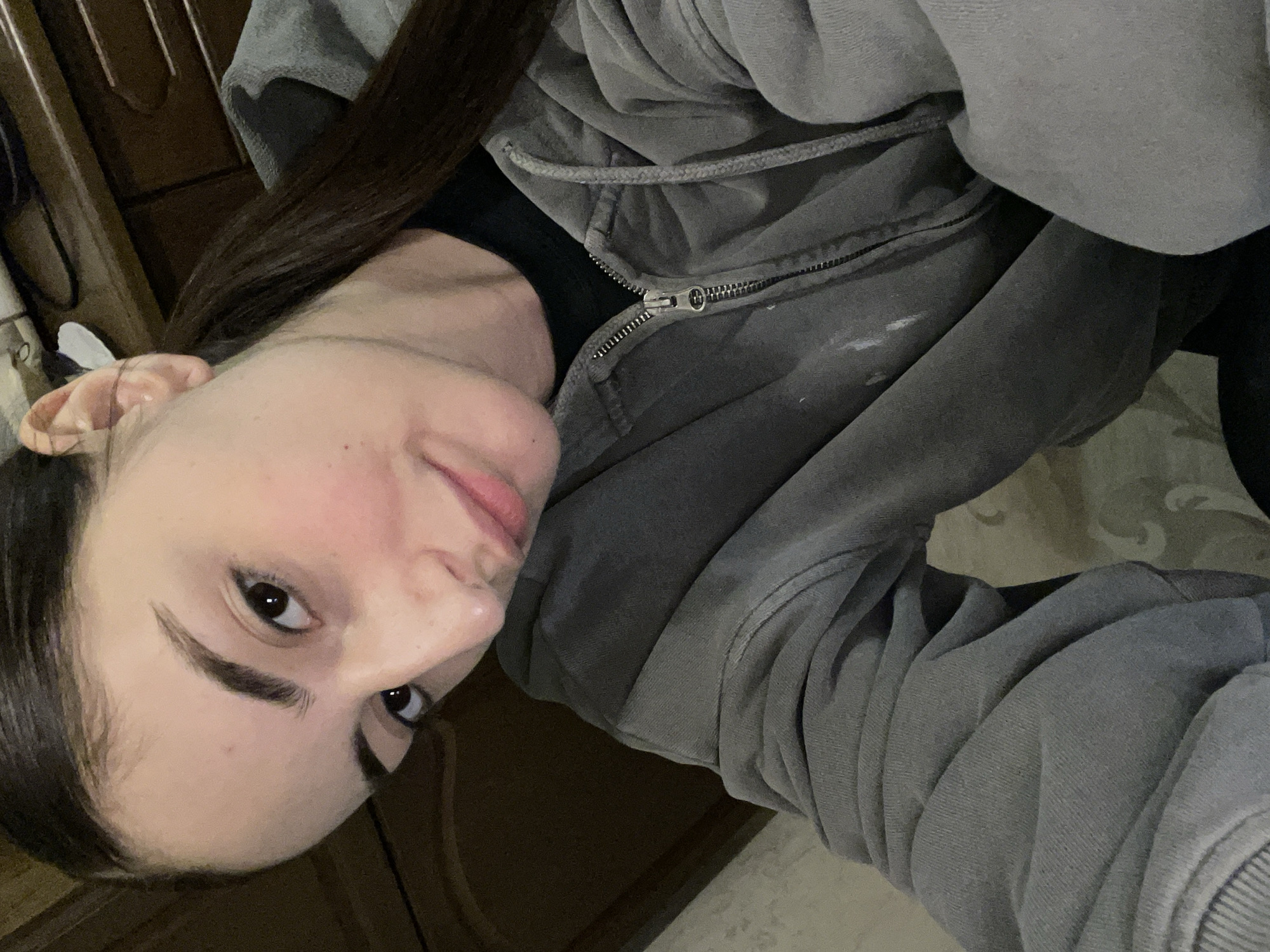 The image depicts a close-up, upward-angle selfie of a young female indoors. She has dark brown hair pulled back into a ponytail, dark eyes, and prominent eyebrows, with a light complexion showing a slight natural flush on her cheeks. Her expression is neutral with a subtle hint of a smile as she looks directly at the camera. She is wearing a grey, full-zip hoodie, which appears worn and has light-colored marks or stains on the left front side near the zipper, over a black top. The background is an interior setting with dark wooden furniture or paneling visible on the left and upper right. A light-colored wall or wallpaper with a faint, repeating pattern is present on the right and bottom sides. A small white object and a dark cable are partially visible in the upper right background, behind her head. The specific location within Moscow, Russia, cannot be determined from the visual information present in the image.