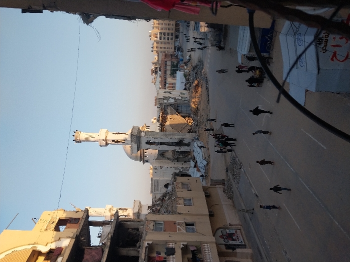 This image depicts an urban landscape in Khan Yunis, Palestinian Territory, exhibiting widespread structural damage. Viewed from an elevated perspective, a central feature in the mid-ground is a large mosque with a partially damaged white minaret and dome. Adjacent multi-story buildings, particularly a yellow-toned structure on the left and another on the right, show severe destruction, including partial collapses, exposed internal elements, and missing sections. Extensive rubble and debris are visible on the ground, especially around the mosque and damaged buildings. A paved street with white lane markings runs through the foreground and mid-ground, containing scattered debris along its edges. Approximately 30-40 individuals, appearing to be adults and children, are present, walking along the street and through the areas of debris. A white tent-like structure with Arabic script is visible in the lower right foreground, beside which a bicycle rests. The scene is illuminated by natural light under a clear blue sky, suggesting daytime.
