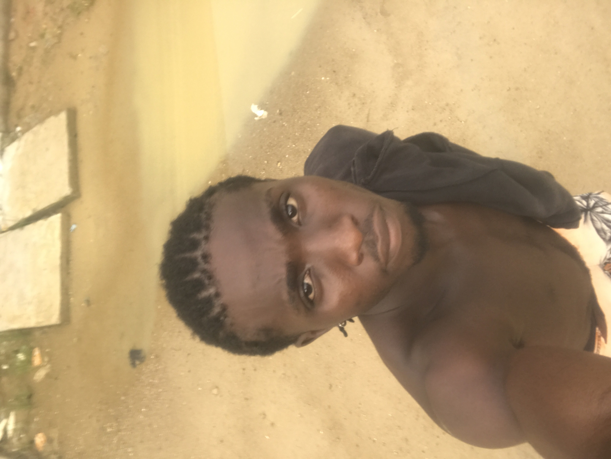 A high-angle, medium shot captures a man with dark skin and braided hair taking a selfie outdoors. He is shirtless, with a dark garment draped over his shoulder. His gaze is directed towards the camera, and his facial expression is neutral. The background consists of sandy ground with some debris, a body of murky water, and two large, flat concrete slabs. The lighting suggests an overcast day.