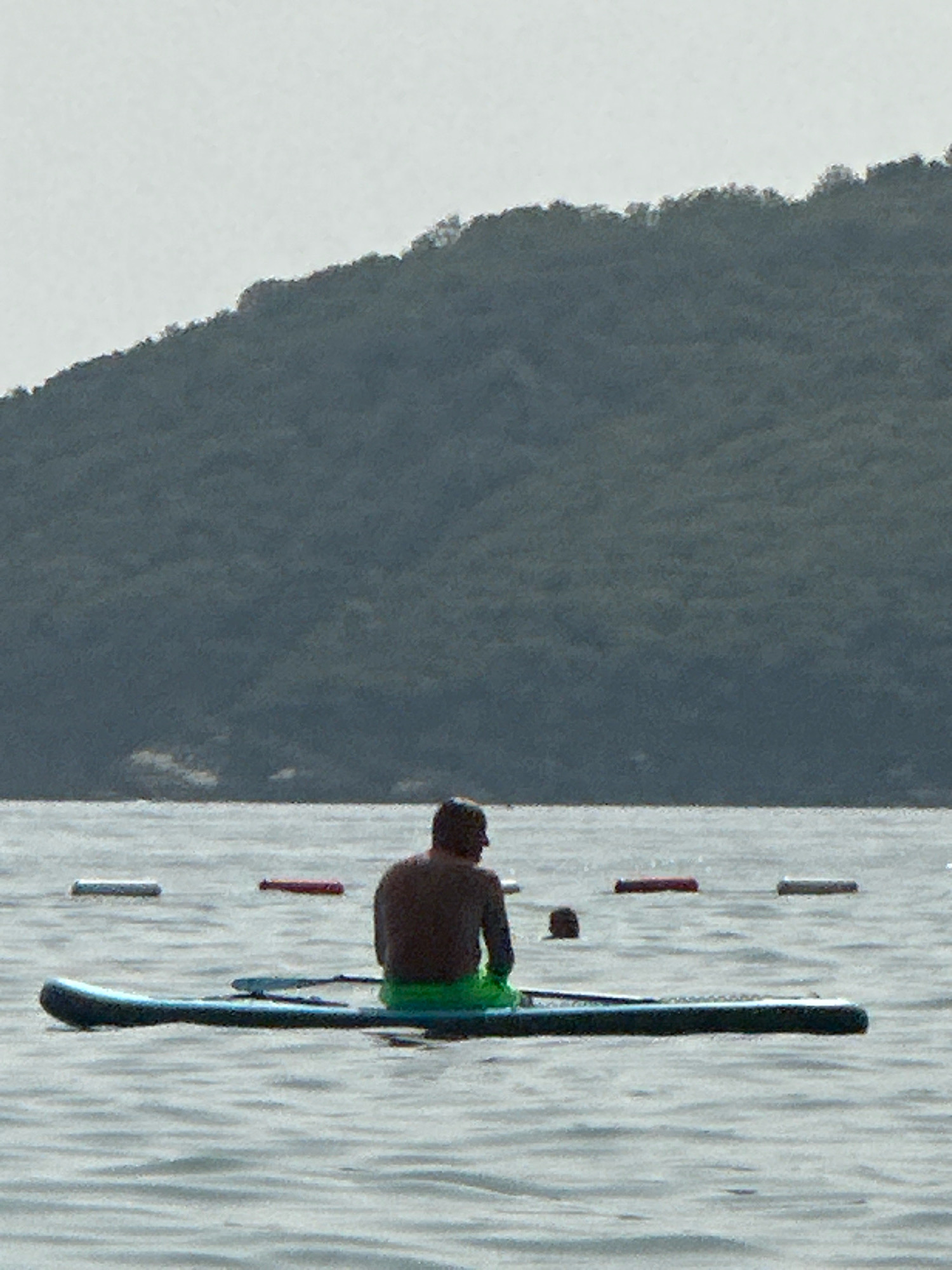 A person sits on a paddleboard in calm water with a dark, tree-covered hill in the background. The person is facing away from the camera, wearing a dark top and green shorts. Two buoys, one white and one red, are visible in the water to the left and center of the frame, respectively. Another person's head is visible in the water to the right of the buoys. The sky is overcast and light grey. The scene appears to be a serene outdoor location, possibly a lake or a calm sea bay. The location in Boreti, Montenegro, is likely a coastal area known for its natural beauty and water activities.