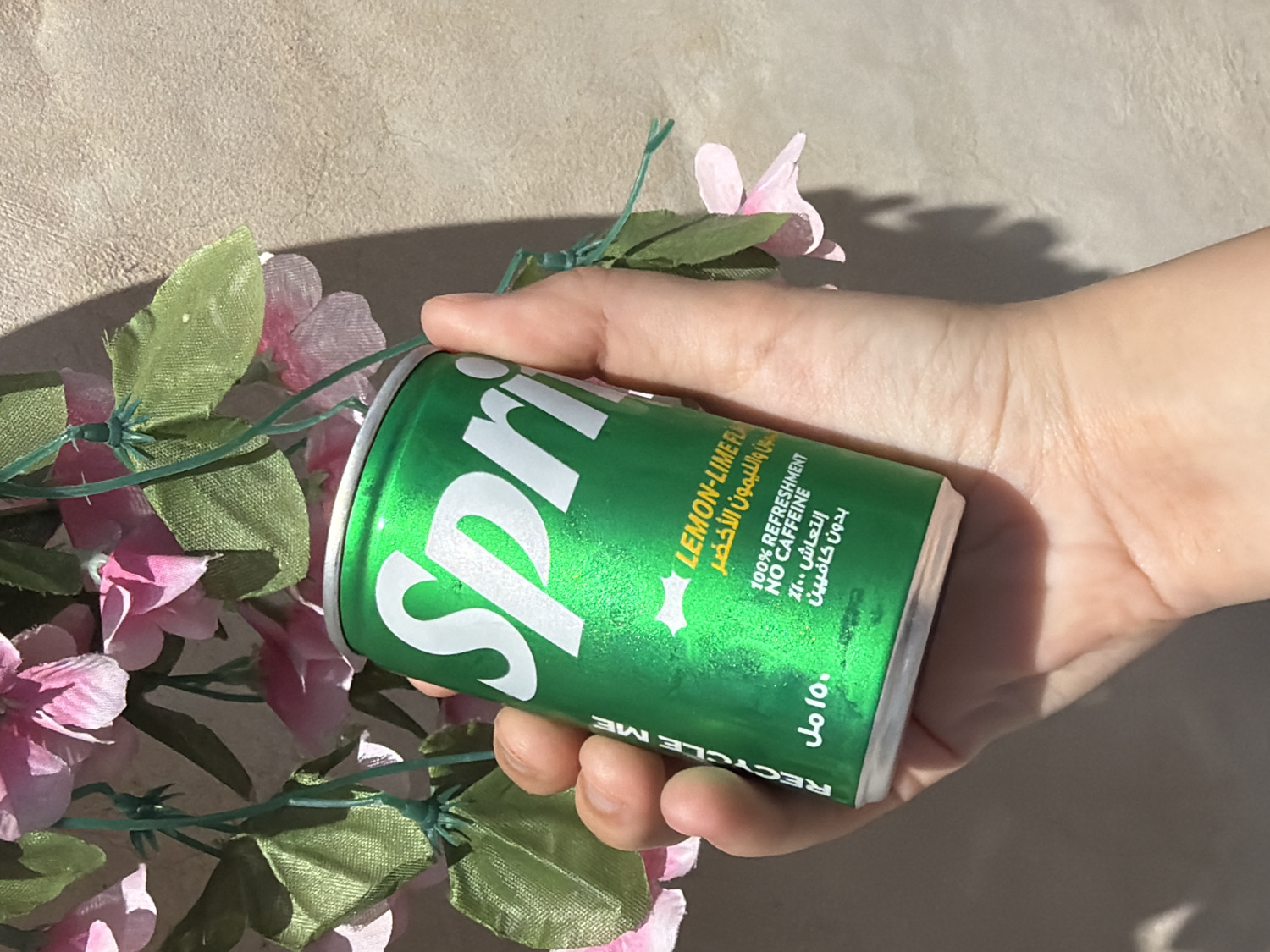 A person's hand is prominently featured, holding a small, vibrant green Sprite can. The can is angled to display its label, which includes text in both English and Arabic. Visible English text reads "Spri" (part of Sprite), "LEMON-LIME FLAVOR," "NO CAFFEINE," "100% REFRESHMENT," and "RECYCLE ME." Corresponding Arabic text includes "١٥٠ مل" (150 ml), "الليمون الأخضر" (green lemon), "انتعاش ١٠٠٪" (100% refreshment), and "بدون كافيين" (no caffeine), consistent with products available in Tel-Aviv, Israel.
The scene is set against a light, textured background, possibly a wall or tabletop, and is adorned with artificial pink flowers and green leaves, connected by visible green wire stems. These floral elements are positioned mostly to the left and beneath the can, adding a decorative touch.
The image is captured in bright natural light, evident from the strong, distinct shadows cast by the hand and the flowers on the background surface, suggesting it was taken during the day. The overall impression is that of a casual product shot, highlighting the beverage in an aesthetically pleasing floral setting.