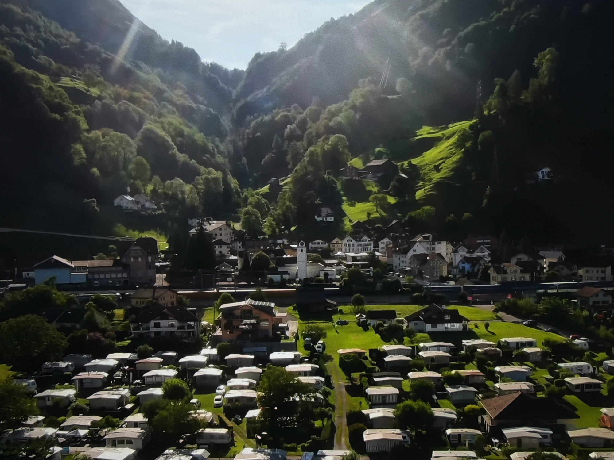 The image shows a panoramic view of a valley town nestled between steep, tree-covered mountains. The sun is shining, casting bright rays through the foliage and illuminating patches of green on the hillsides. In the foreground, there is a large mobile home park with numerous white and light-colored trailers arranged in rows. Beyond the trailer park, there is a town with various buildings, including houses, a church with a tall white steeple, and what appears to be a school or administrative building. A train track runs through the town, and a train is visible moving along it. The overall scene depicts a rural or semi-rural setting with a mix of residential areas and natural landscapes.
