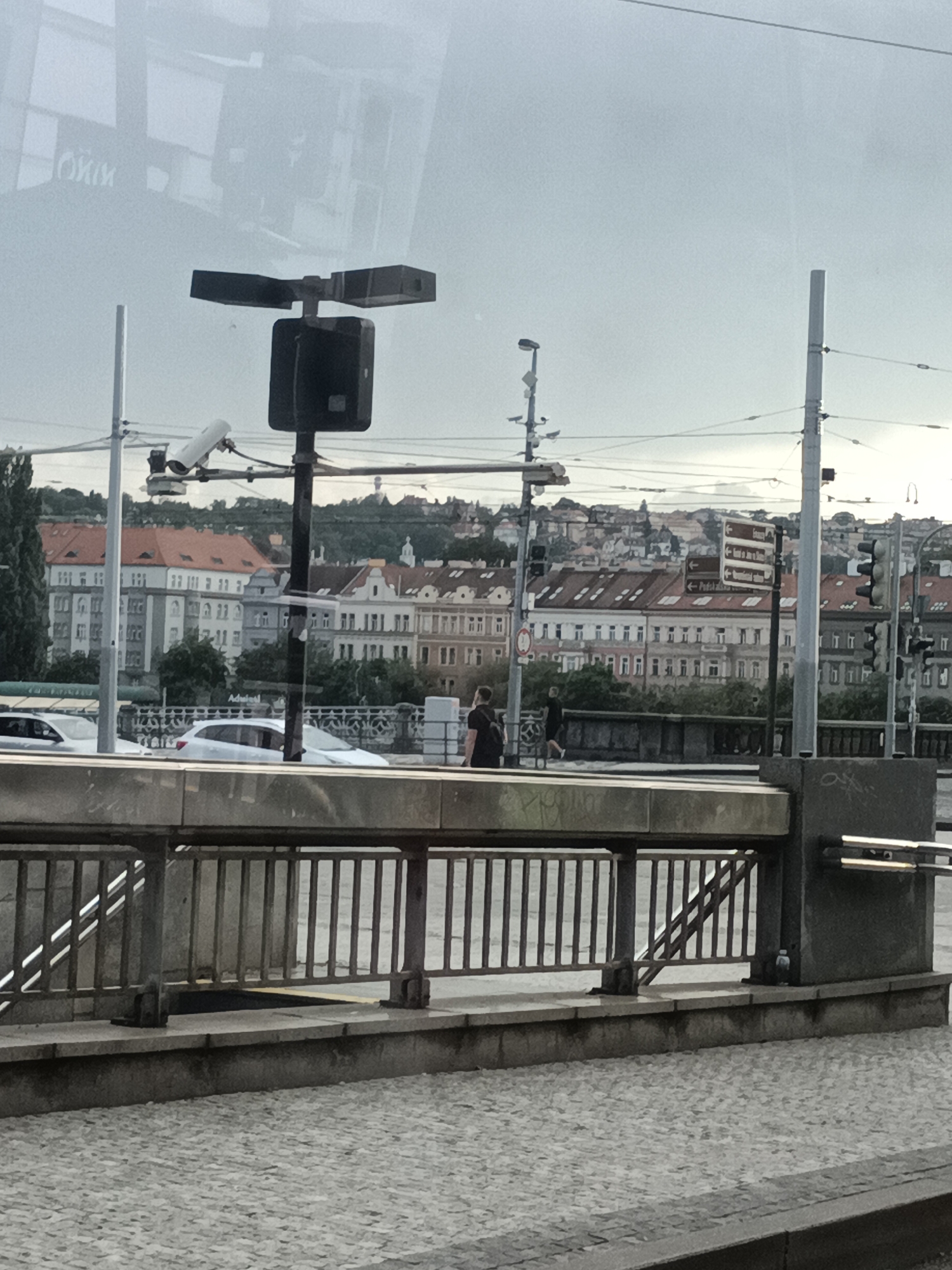 The image depicts a street scene in Prague, Czechia, with a focus on a pedestrian walkway or bridge area. In the foreground, there is a metal railing with a stone base, characteristic of urban infrastructure. Behind the railing, a cobblestone street is visible, with a few cars, including a white hatchback, parked or driving. Further in the background, a dense cluster of European-style buildings with red-tiled roofs and multi-story facades line a hill. Various streetlights and traffic signals are present, indicating an active urban environment. A person in dark clothing is walking away from the camera on the right side of the image. Reflections on a glass surface, likely from a window or tram, obscure parts of the scene, showing upside-down text and architectural elements. A security camera is mounted on a pole to the left of the frame, overlooking the street. The overall atmosphere is overcast, suggesting a cloudy day.