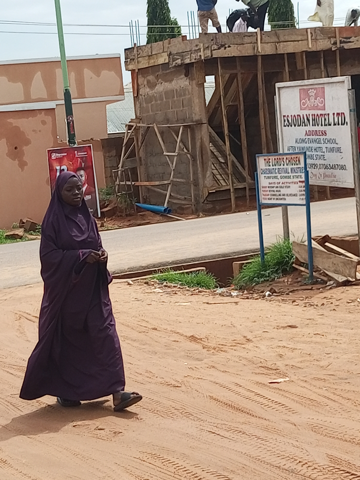 A woman dressed in a dark purple hijab and abaya walks across a dusty dirt road in Tempure, Nigeria. She is walking from left to right in the frame and appears to be looking down. Behind her, a building is under construction. A sign with "ESJODAN HOTEL LTD." is visible on the right side of the frame, along with another sign that reads "THE LORD'S CHOSEN CHARISMATIC REVIVAL MINISTRIES". The environment is arid and dusty, with some green vegetation growing near the signs. Tire tracks are visible in the dirt road.