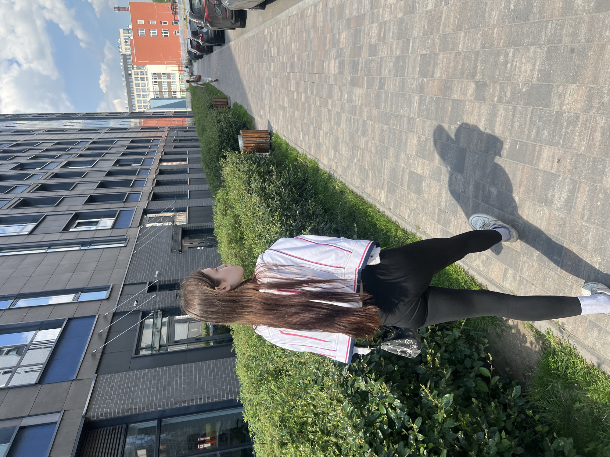The image captures a young woman standing on a paved sidewalk in a modern urban environment in Saint Petersburg, Russia, on a bright, sunny day. She is seen in profile, looking towards the right, with long brown hair cascading down her back. She is dressed in a white jacket with red and blue stripes, black leggings, white socks, and white sneakers, and carries a small black purse with white doodles.
To her left, a tall, contemporary dark grey apartment building with dark brickwork and large windows dominates the frame. A line of green bushes and grass separates the building from the patterned grey paving stones of the sidewalk. Further down the sidewalk, small wooden benches are visible among the foliage.
To her right, the paved path extends into the distance, flanked by parked cars and another person walking further away. In the background, a prominent reddish-brown building with numerous windows stands against a clear blue sky dotted with white clouds. Strong shadows of the woman and other objects are cast onto the ground, indicating bright sunshine. A partially obscured red sign with white text is visible on the dark grey building. The scene depicts a serene moment in a well-maintained urban setting.