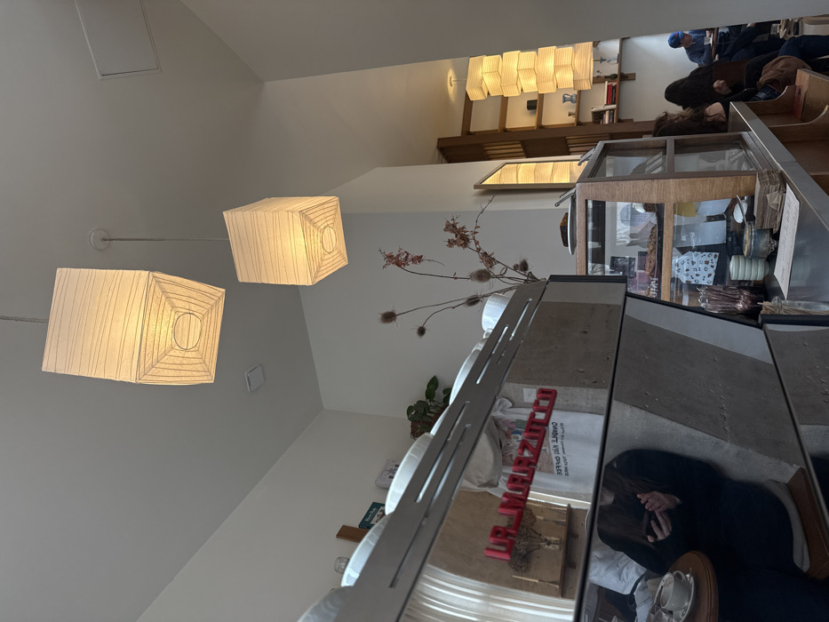 The image displays the interior of a minimalist coffee shop, identified as Oud Paris, located at 20 Rue de Champollion, 75005 Paris, France. The scene is brightly lit by natural light and several warm-toned, rectangular paper lantern pendant lights suspended from a high white ceiling.
In the foreground, a large, reflective stainless steel La Marzocco espresso machine is prominent on the left. Its surface mirrors a person with long dark hair, dressed in a dark top, seated on a light-colored stool, actively viewing a smartphone held in both hands. A white cup and saucer are positioned on a small round wooden table next to the reflected individual. White ceramic cups are stacked neatly on top of the espresso machine.
Towards the center, behind the espresso machine, dried thistles or similar plants are arranged in a vase on a counter. Further right, a wooden display case with glass panels holds various baked goods. A rectangular mirror hangs on the wall, reflecting interior elements.
The background to the right reveals a seating area with patrons. At least three individuals are partially visible; one person with dark hair is seated with their back towards the camera, and another wearing a blue cap is seated further back at a table. A tall, vertically stacked series of rectangular paper lantern lights illuminates this area, adjacent to a wall-mounted wooden shelving unit. The walls are painted white, contributing to a clean, open aesthetic.