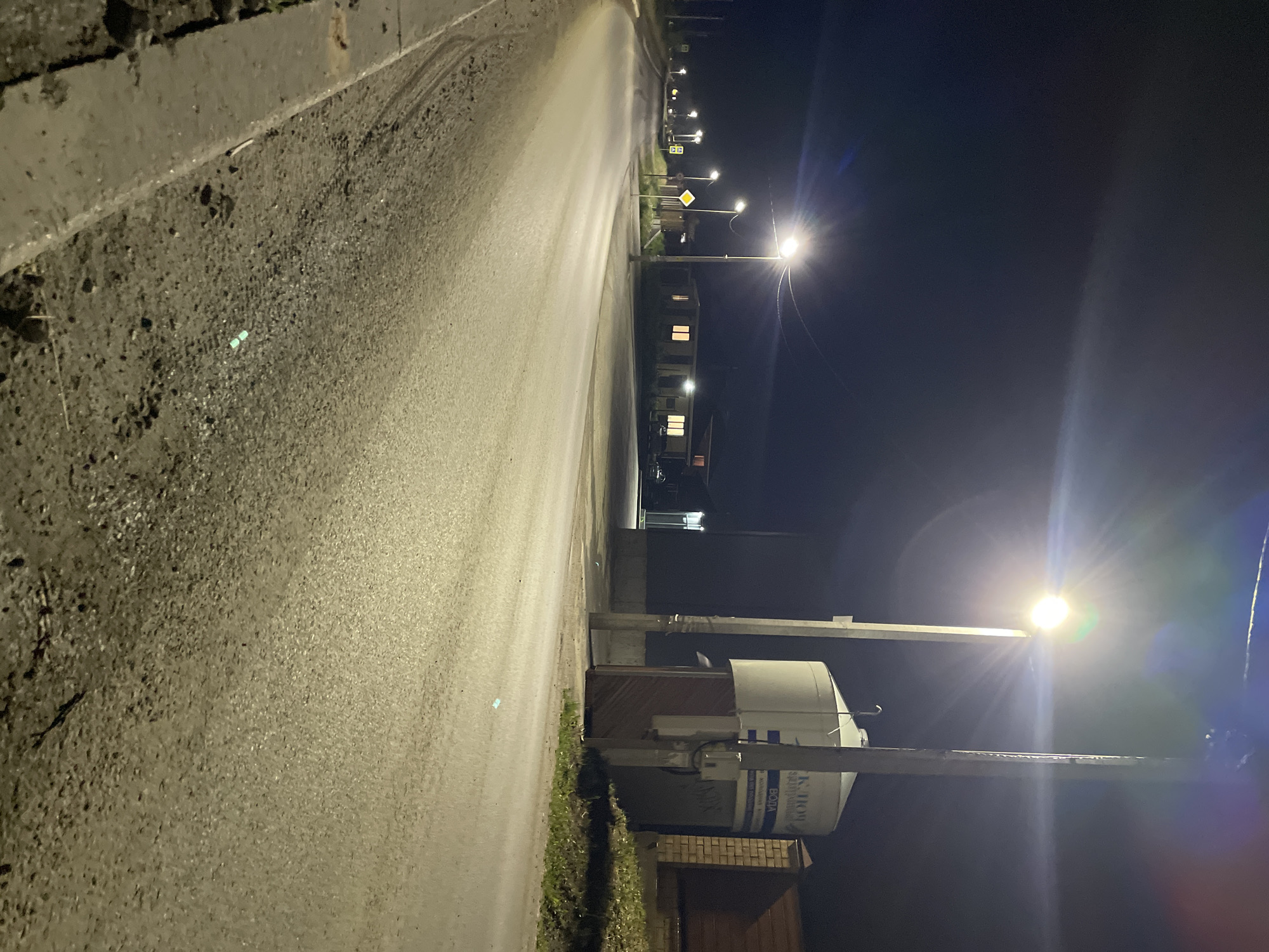 A nighttime scene depicts a street in Эстачи, Russia, illuminated by streetlights. The asphalt road stretches into the distance, with a sidewalk to the left. Buildings are visible along the right side of the road, some with lit windows. A utility structure with a cylindrical tank is visible in the foreground.