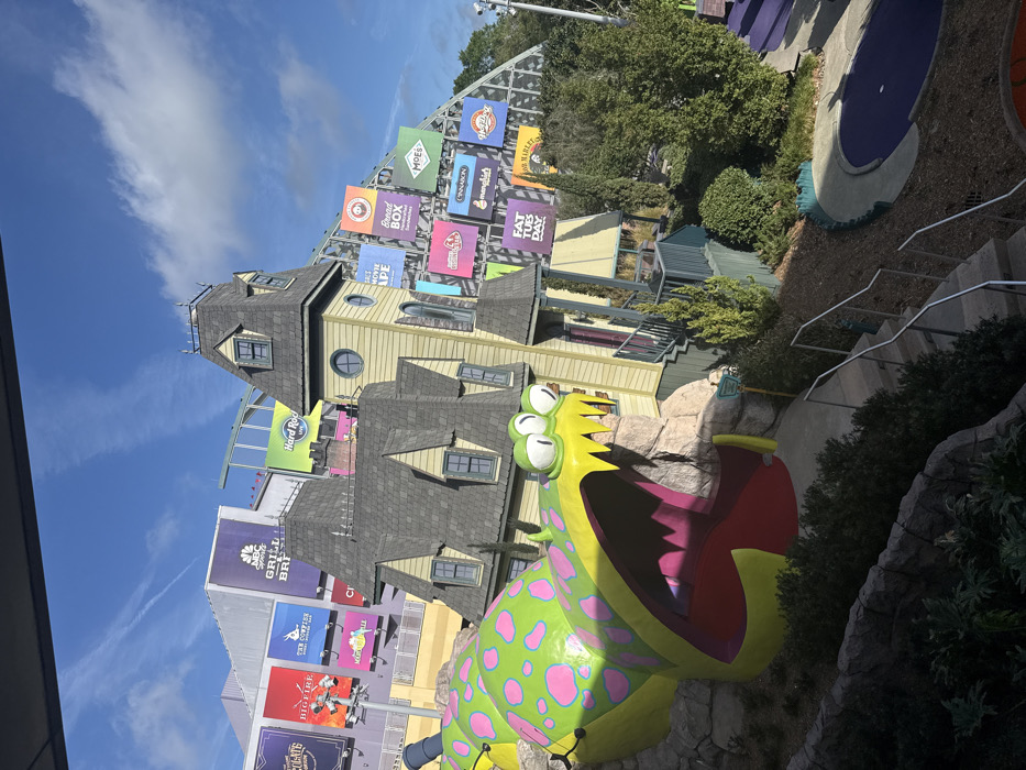 This image depicts a themed outdoor entertainment area within Universal Orlando Resort in Orlando, Florida, USA, under a clear blue sky with sparse white clouds.
The central feature is a large, brightly colored sculpture of a monster, identified as "Fluffy" from the *Despicable Me* franchise. It is predominantly lime green with large pink polka dots, featuring three prominent eyes and a wide-open mouth with yellow, pointed teeth revealing a red interior. The monster emerges from a brown, rocky base. To its right stands a two-story, light yellow house with dark grey peaked roofs and dormer windows, designed in a deliberately skewed or crooked architectural style characteristic of Gru's house from *Despicable Me*.
In the background, a large building complex is visible, adorned with numerous advertising billboards for various Universal Orlando dining and entertainment establishments. These signs include "NBC Sports Grill & Brew," "Hard Rock Live," "The Toothsome Chocolate Emporium & Savory Feast Kitchen," "Bigfire," "The Cowfish Sushi Burger Bar," and "Margaritaville." Further to the right, another set of brightly colored billboards on a structural framework advertises "Salt & Straw," "Bread Box Sandwiches," "Moe's Southwest Grill," "Red Oven Pizza Bakery," "Cinnabon," "Menchie's Frozen Yogurt," "Hot Dog Hall of Fame," and "Bob Marley – A Tribute to Freedom."
In the foreground and extending towards the lower right, a multi-level concrete staircase with metal handrails descends. Below, part of a purple-colored mini-golf hole is visible, containing a single white golf ball. The area is landscaped with various green trees, shrubs, and bushes. No individuals are present in the scene.