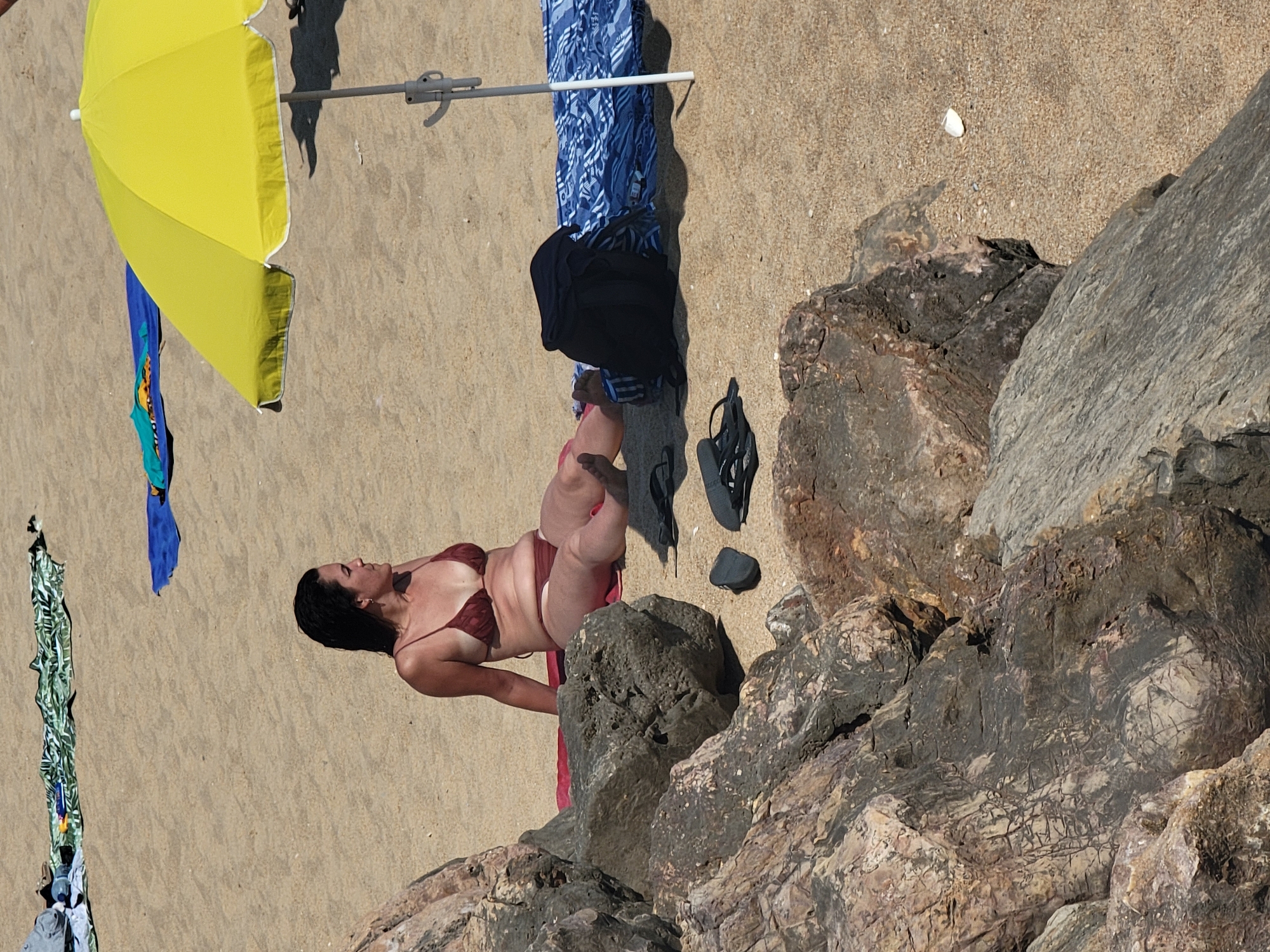 The image depicts a woman sunbathing on a sandy beach in Quarteira, Portugal. She is lying on her back, wearing a maroon bikini and her eyes are closed, suggesting she is relaxed and enjoying the sunshine. A yellow umbrella is partially visible to her left, providing shade. Various items of clothing are scattered on the sand around her, including a blue and white patterned fabric, a black t-shirt, and a green and white patterned fabric. Black flip-flops are also present near her feet. The beach is a mix of sand and rocks, with larger rocks forming a prominent feature in the foreground. The scene appears to be set during the daytime, with bright sunlight casting shadows. There is no visible text in the image.