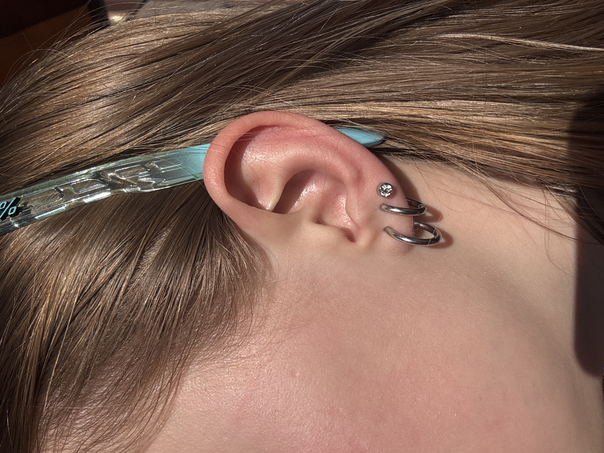 A close-up photograph shows a person's left ear with multiple piercings. The uppermost piercing is a small stud with a clear gemstone. Below it are two silver-colored hoop earrings, creating a stacked effect. The ear is pinkish in color, contrasting with the silver jewelry. The person's light brown hair is visible around the ear. A light blue hair clip with illegible silver characters is partially visible behind the ear. The skin tone appears fair and without blemishes. The background is out of focus, suggesting a shallow depth of field. No elements suggest a location within Gory, Russia.