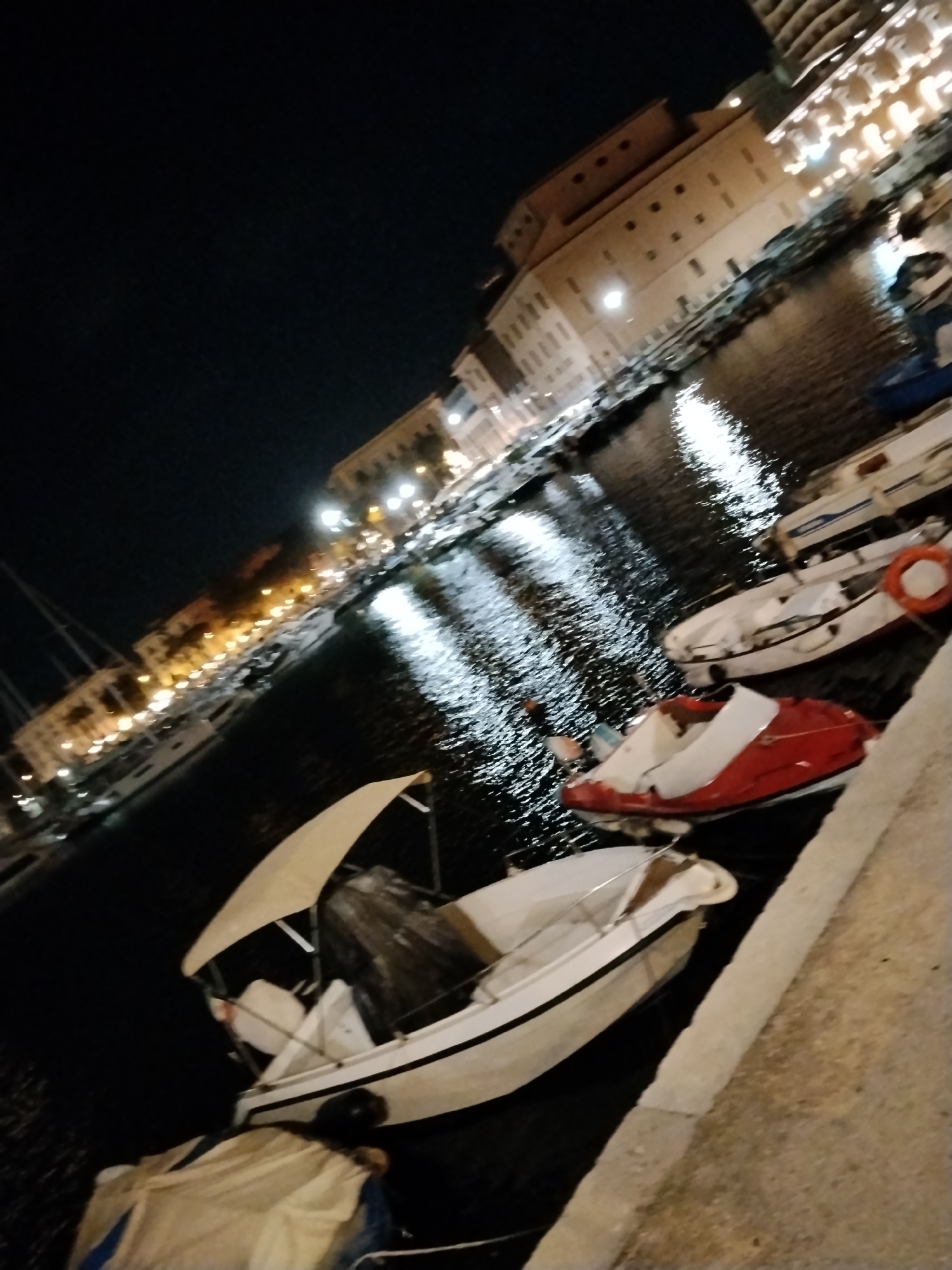 At night, in the city of Bari, Italy, a fleet of boats is docked along a waterfront. The buildings lining the harbor are illuminated, casting reflections on the dark water. The boats are of various sizes and types, including a white boat with a canopy, a red and white speed boat, and several other smaller vessels. One of the boats has a lifebuoy attached to it. The scene is tranquil, with the lights of the city creating a warm ambiance. The perspective of the image is slightly tilted, adding a dynamic quality to the composition.