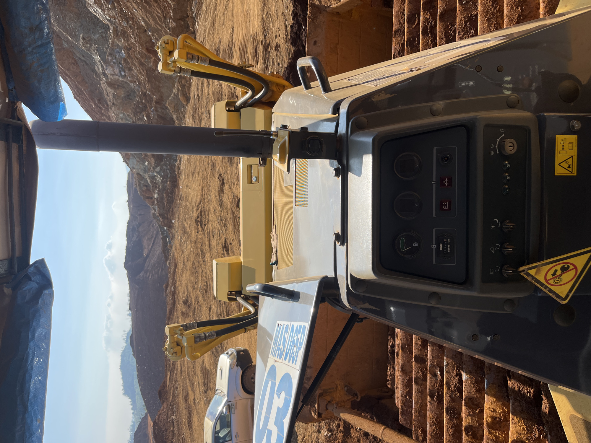 This is a first-person perspective photograph taken from the operator's seat of a bulldozer at a construction site. The bulldozer's controls and dashboard are in the foreground. Behind the bulldozer, a large excavation area with reddish-brown earth and rock formations is visible. The sky is blue with scattered white clouds. In the distance, a silver pickup truck is parked on the site. The overall environment suggests an active mining or large-scale earthmoving operation. The location is identified as Unknown, Indonesia, although no specific landmarks within the city are visible.