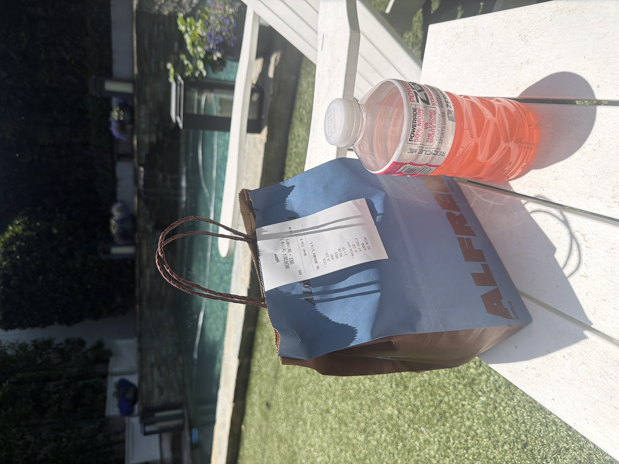 The image shows a Powerade Zero sports drink bottle and an Alfred Coffee blue paper bag on a white surface, likely an outdoor table, with a receipt sticking out of the bag. The receipt indicates the order was from "UBEREDBC4 Tallia S." and was delivered via Uber. The order includes a latte, iced drinks, honey, breve, regular espresso, matcha latte, sugar-free vanilla, almond milk, and regular matcha. The bag displays the Alfred Coffee logo, with the phrase "Alfred.LA" printed beneath. The background features greenery, including a lawn, shrubs, and trees, suggesting an outdoor patio or garden setting. The location is likely in Los Angeles, United States of America, based on the receipt information.
