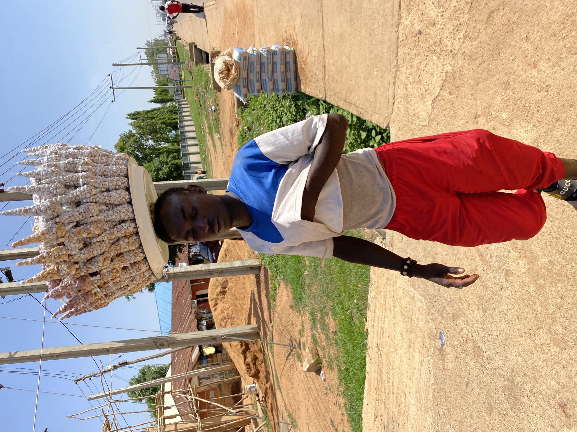 A man wearing red pants, a gray and blue shirt, and black shoes stands on a concrete surface in Bukuru, Nigeria. He balances a large, flat tray on his head, loaded with numerous small bags of what appears to be peanuts. A bracelet adorns his left wrist. He looks directly at the camera, with his hand open to the side. In the background, there are buildings, sand piles, utility poles, and a street. There are also bags of cement beside the road. Another person is walking in the distance. The sky is light blue.
