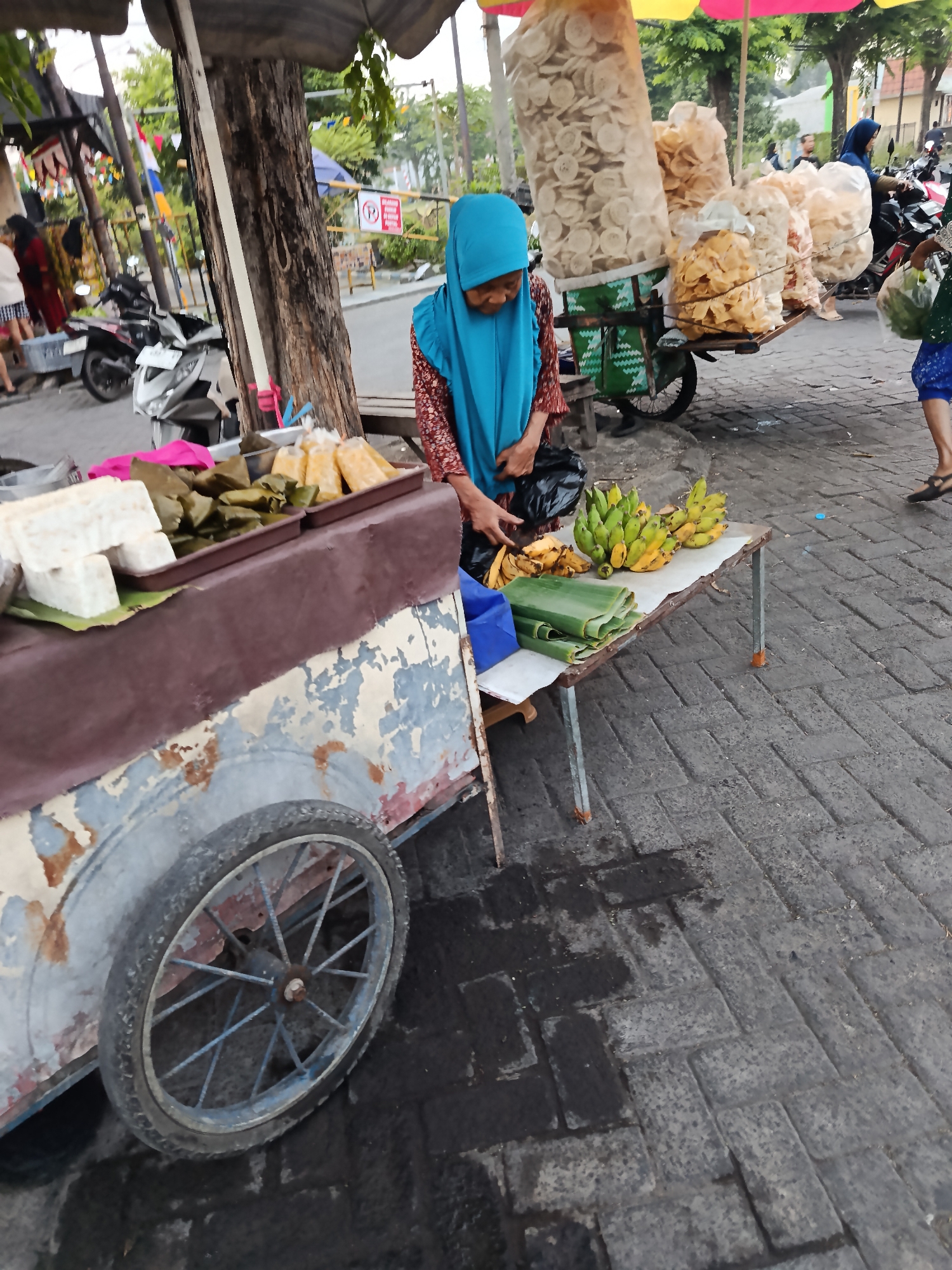 A street vendor in a blue hijab stands behind a cart laden with goods in Keboansikep, Indonesia. The cart, painted in peeling shades of rust and pale blue, has a single spoked wheel. On the cart's surface are blocks of white material, possibly tofu, wrapped packages in banana leaves, and a tray of yellow, individually wrapped items. To the right of the cart, a metal table holds several bunches of bananas and more wrapped items. Behind the vendor, large bags of what appear to be crackers are displayed on a rack, some under a colorful striped umbrella. The ground is paved with dark interlocking bricks. In the background, motorcycles are parked, and other pedestrians are visible. The scene depicts a bustling market environment.