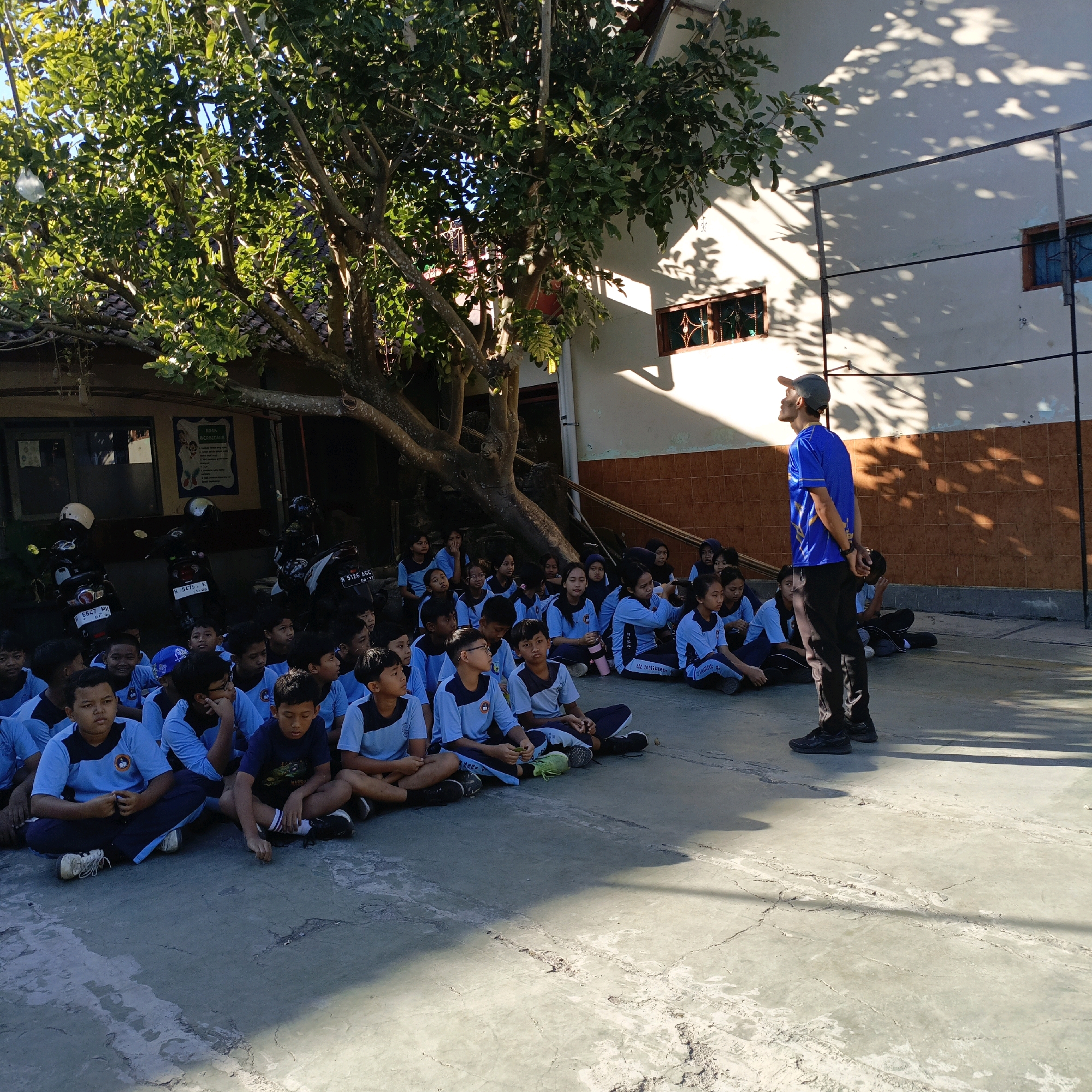In Salatiga, Indonesia, a group of students, likely elementary or middle school age, are sitting cross-legged on a concrete ground in front of a building. They are wearing matching light blue uniforms with dark blue accents. A man in a blue t-shirt and black pants stands facing the students, holding a cap in his hands, and appears to be addressing them. The scene is outdoors, with a large tree casting dappled shadows on the ground and the building's wall. The building has a tiled roof and windows with decorative grilles. Several motorcycles are parked behind the students, suggesting a school or community gathering. The sunlight indicates it is daytime.