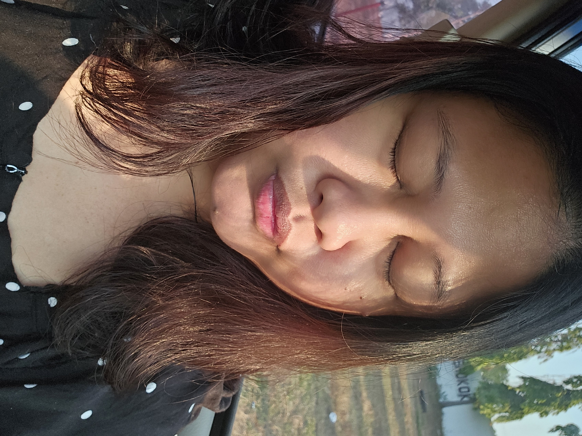 A woman is captured in a close-up shot, her eyes closed and her lips pursed as if puckering. She is seated in a vehicle, with the edge of a car window visible on the left side of the frame. Her dark, straight hair frames her face, with strands falling onto her shoulders and chest. She is wearing a black top adorned with white polka dots. The lighting suggests it is daytime, with sunlight casting a warm glow on her skin and highlighting the texture of her hair. In the background, out of focus, a fence with text that appears to read "BOKOK" is visible, along with trees and a clear sky, indicating an outdoor urban or semi-urban environment. The overall impression is of a candid portrait taken in natural light.