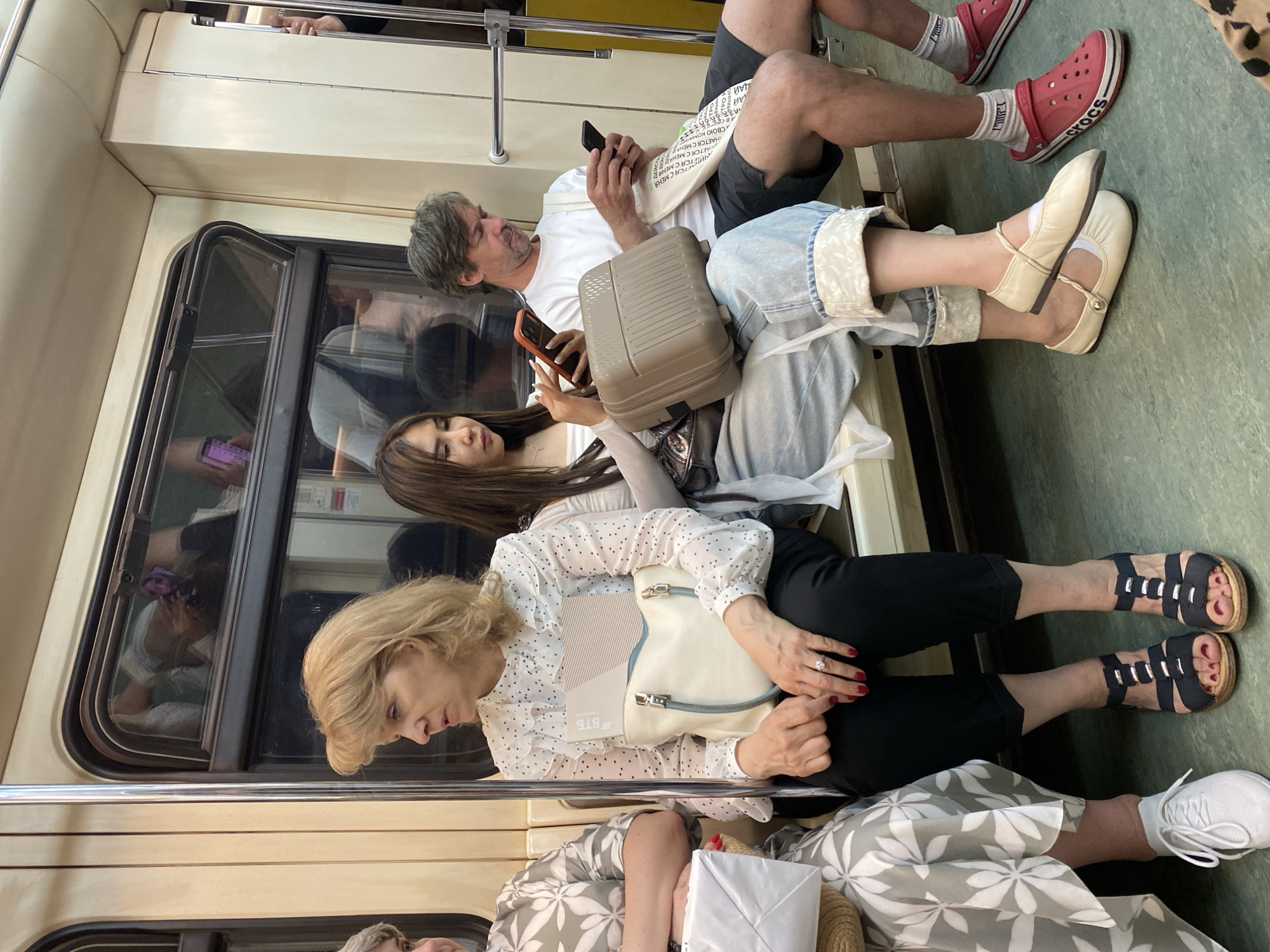 The image shows a scene inside a train car, possibly in Moscow, Russia, indicated by the Cyrillic alphabet on a white bag. Several passengers are seated. A man in a white shirt is looking at his phone, holding a bag with Cyrillic text and a small tan suitcase on his lap. Next to him, a young woman is also looking at her phone. A woman with blonde hair sits below them, holding a white purse and wearing a polka dot blouse. An older woman wearing sandals is seated to the left. Crocs and white sneakers are also visible. The passengers seem occupied with their devices during the commute. The environment is the interior of a train car with windows and interior seating.