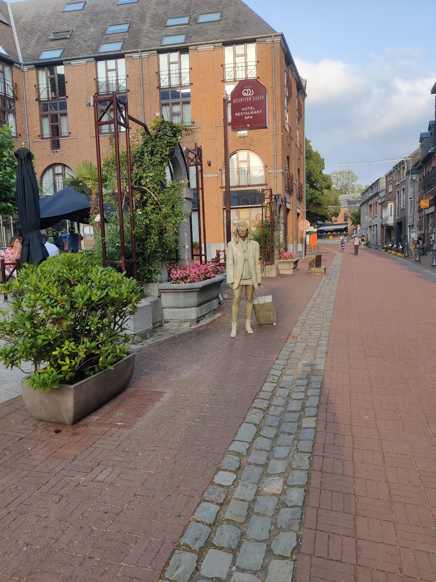 The image captures a street scene in Marche-en-Famenne, Belgium. A paved walkway, constructed with red bricks and a stone border, runs through the frame. On the left side of the walkway, a bronze statue of a woman in a jacket and skirt, holding a suitcase, stands prominently. Behind the statue, a large potted plant with green foliage is visible. Further back, a building with red brick facades, featuring numerous windows and balconies, is present. A sign on the building reads "QUARTIER LATIN HOTEL RESTAURANT SPA". Beyond this building, a street with shops and pedestrians extends into the distance. The sky above is partly cloudy.