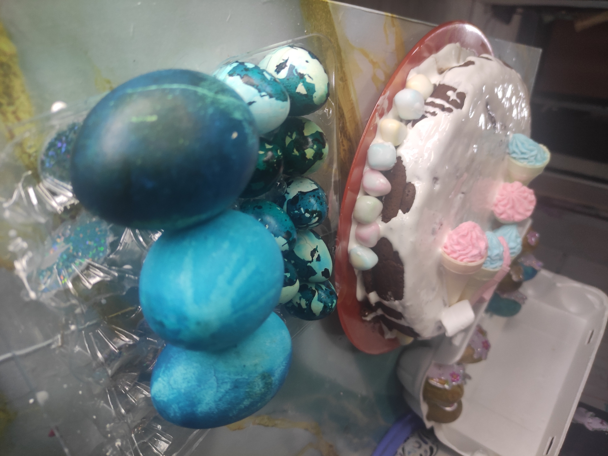 The image shows a table top scene with two main elements: dyed Easter eggs and a decorated cake. The Easter eggs are colored in various shades of blue and green. Some are stacked, while others are in a clear plastic container. The cake is decorated with white frosting, pastel marshmallows, and miniature ice cream cones. The scene appears to be indoors, with a white plastic bin partially visible in the background. There is no information to suggest this is in Chaikovsky, Russia, so I cannot include this in the description.