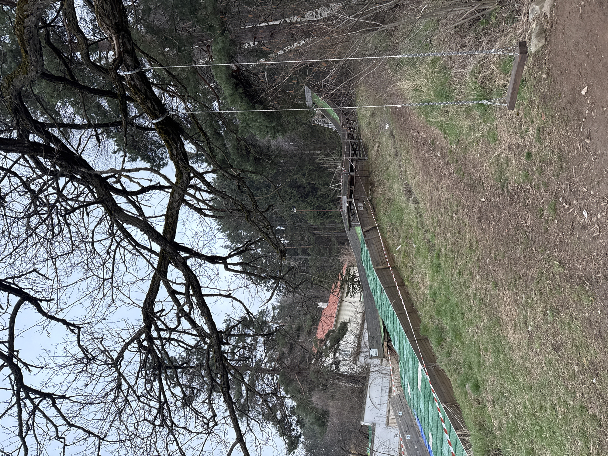 The image depicts a scene in Sofia, Bulgaria, showing a dirt path alongside a fence with green mesh and red and white safety tape. The path is edged with dry grass and some green patches. A series of wooden posts connected by metal chains run alongside the path. Behind the fence are buildings, including one with a red-tiled roof. A ski jumping ramp is visible in the background. A large tree with bare branches dominates the left side of the image, partially obscuring the sky. Other trees, including pines and birches, are visible in the background.