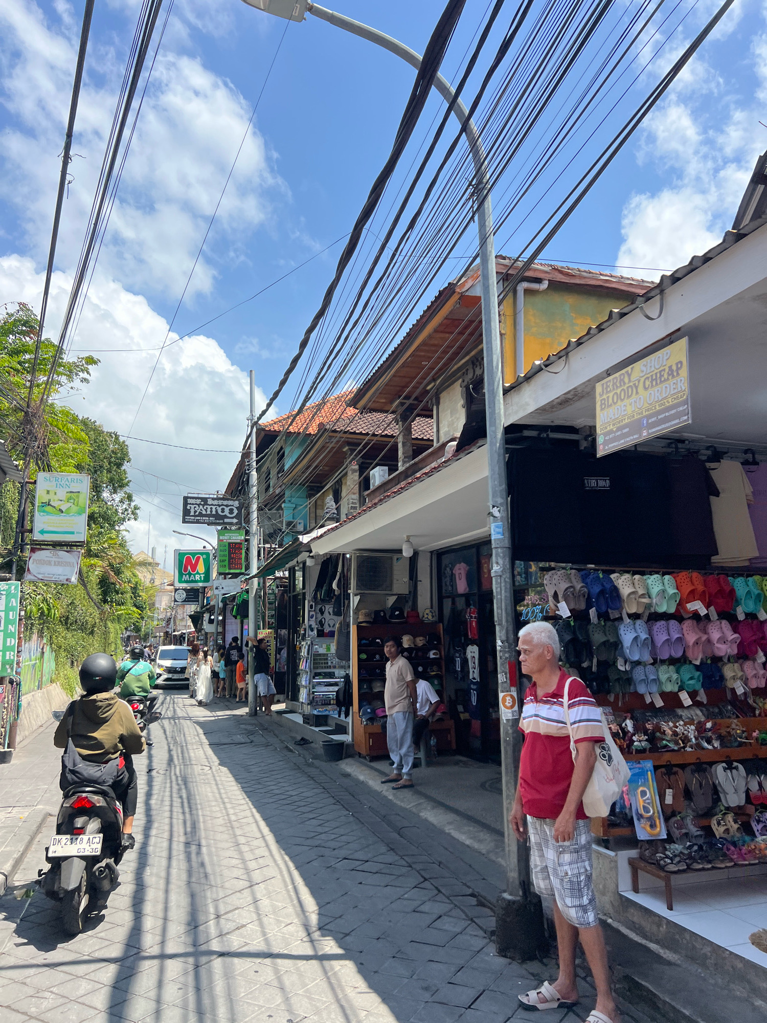 The image shows a street scene in Kuta, Indonesia. A person rides a scooter down a narrow, paved street lined with shops on both sides. Overhead, a tangle of electrical wires and a street lamp are visible against a blue sky with scattered clouds. The shops display clothing and footwear, with a "JERRY SHOP BLOODY CHEAP MADE TO ORDER" sign prominent above one establishment. Several people are walking along the sidewalk, and a white car is parked further down the street. The atmosphere appears to be daytime, with sunlight casting shadows on the ground.