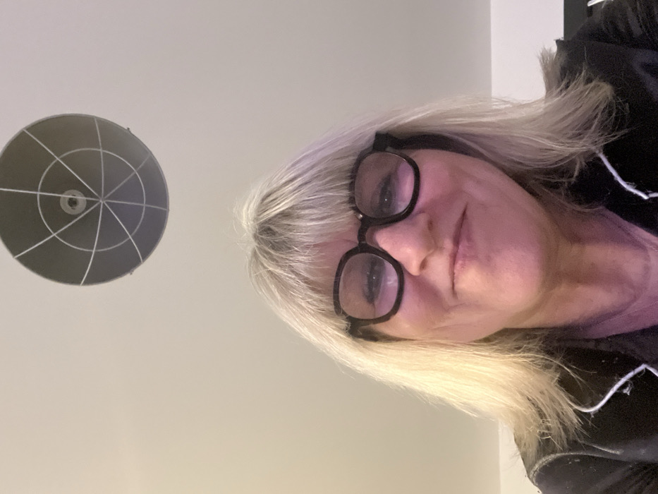 A blonde woman with glasses is angled towards the camera. She wears a black top with white trim. Above her head, on a light-colored ceiling, is a dark, circular light fixture with a geometric pattern of white lines radiating from the center. The woman's face is clear, and her expression is neutral.
